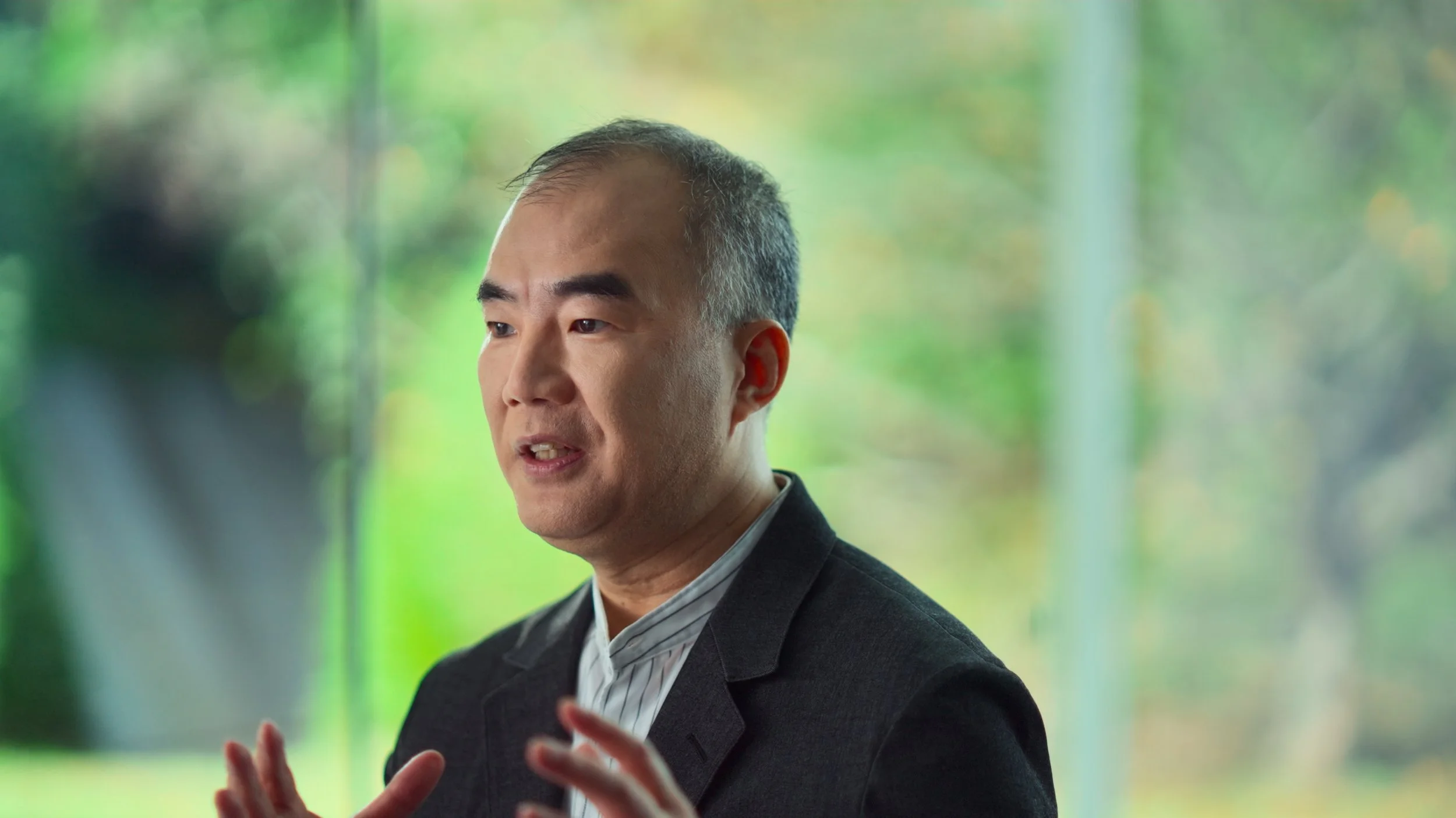 A man with short dark hair and a light complexion speaking, wearing a dark blazer over a striped shirt, with a blurred green outdoor background.