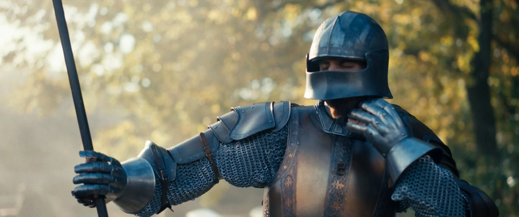 A person dressed as a medieval knight in armor, holding a spear, and adjusting their helmet outdoors with blurred trees in the background.