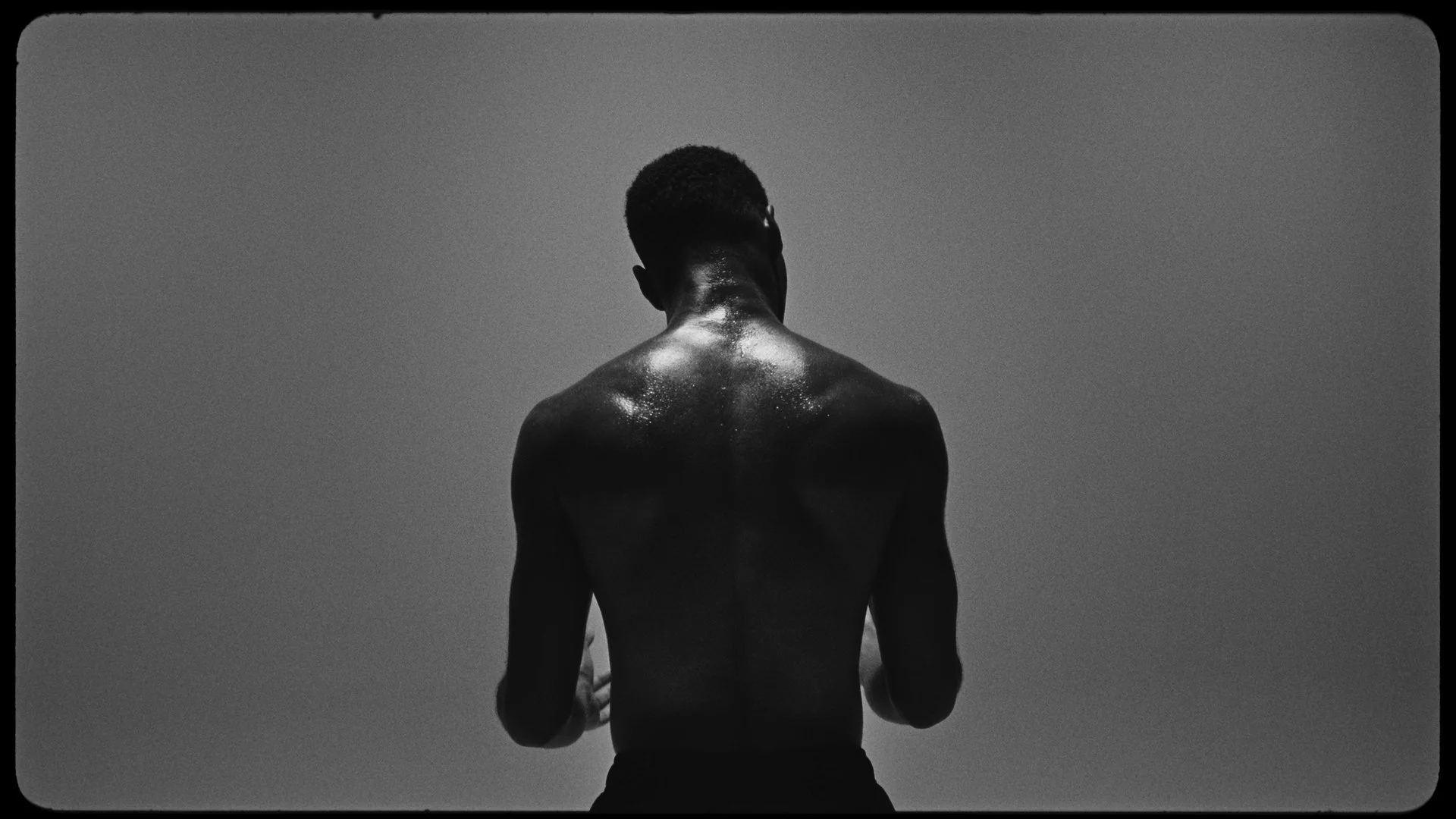Black and white photograph of the back of a shirtless man with short hair, standing outdoors against a clear sky.