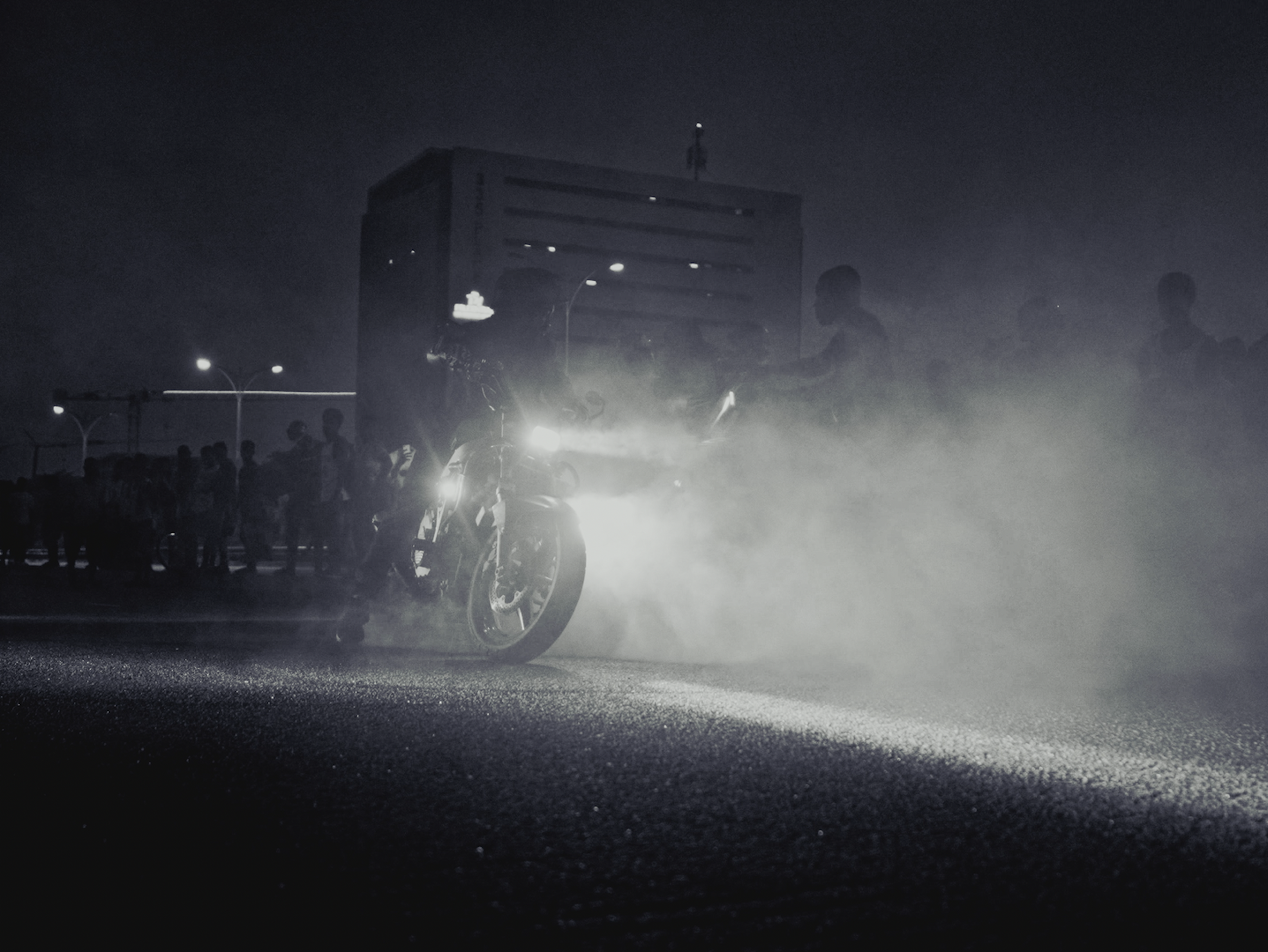 A dark scene with silhouettes of people and a motorcycle, with a building in the background and headlights shining on the ground.