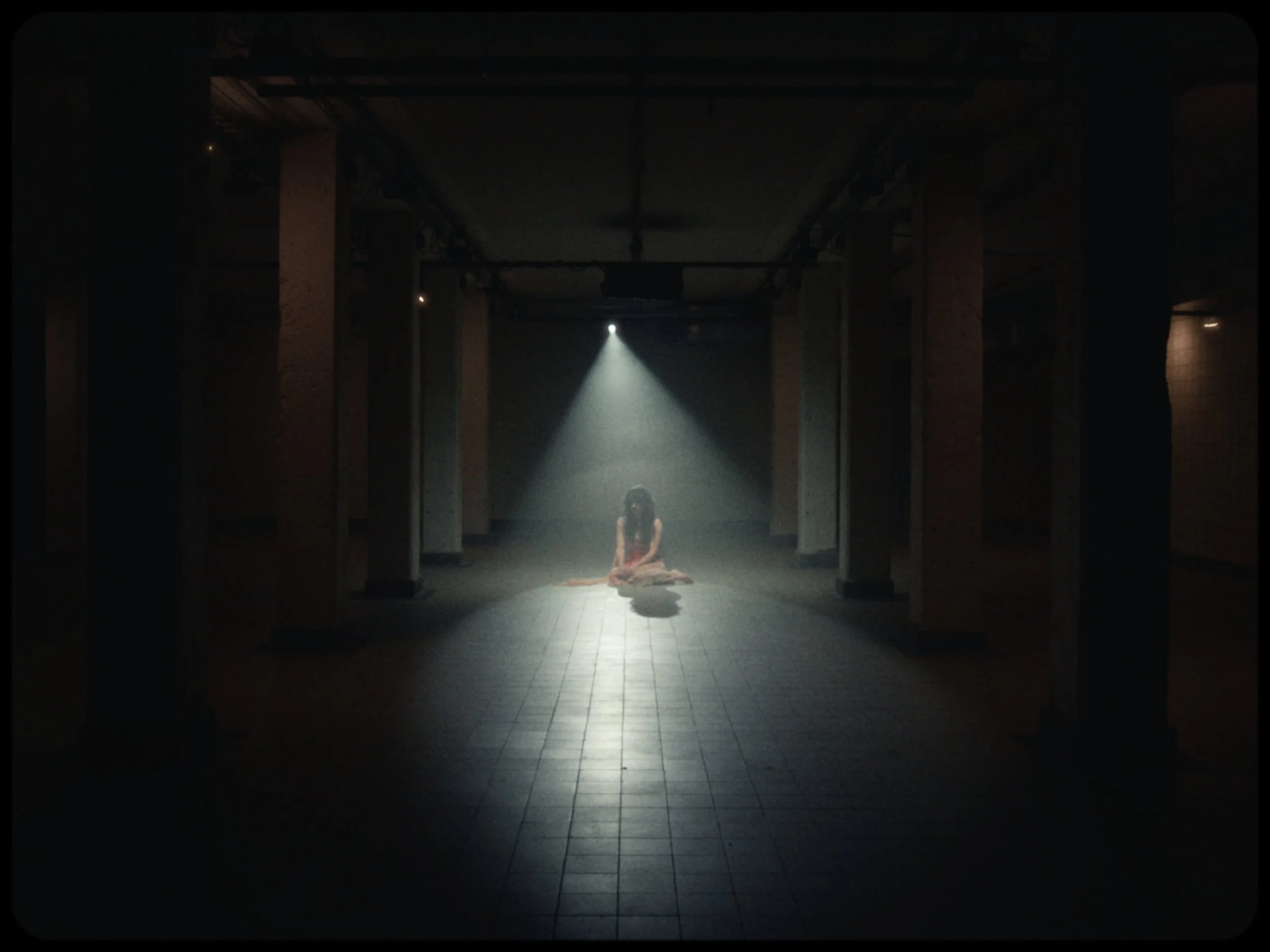 A woman sitting on the floor in a dark, empty corridor illuminated by a single bright light shining from above.