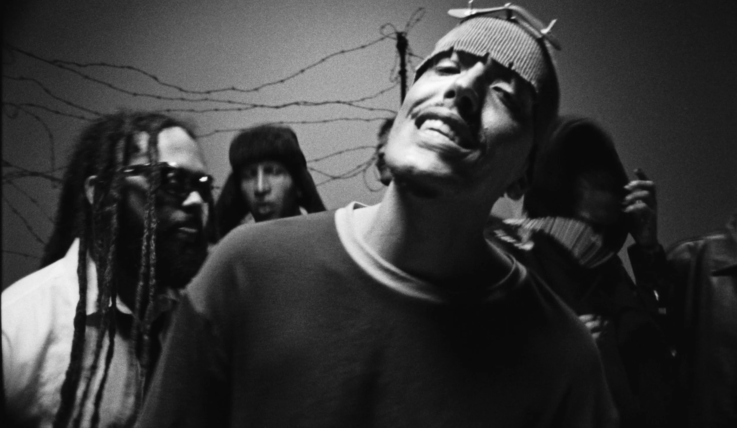 Black and white photo of a man smiling with eyes closed, wearing a beanie, surrounded by people, with barbed wire in the background.
