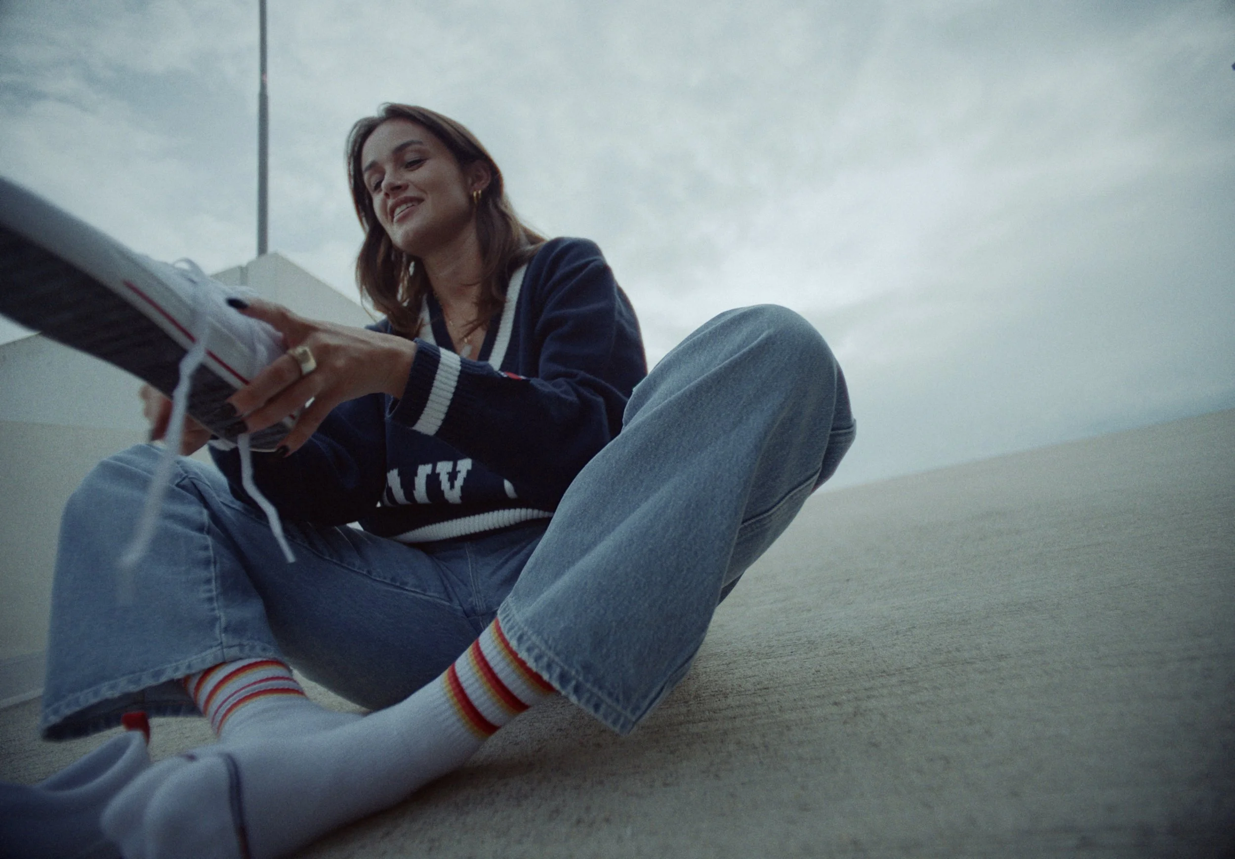 A woman sitting on the ground, smiling and looking down at a magazine she is holding, outdoors on a cloudy day.