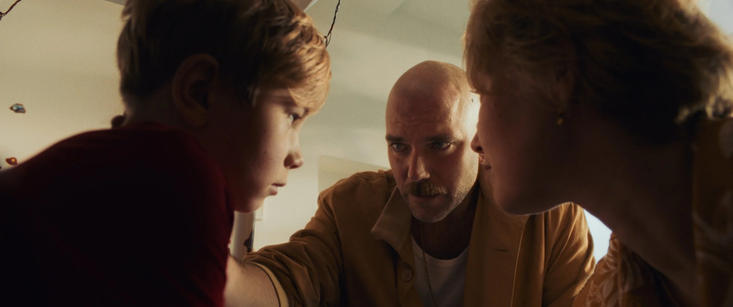 A man with a bald head and a mustache is closely inspecting two children, one boy and one girl, in a room with warm lighting.