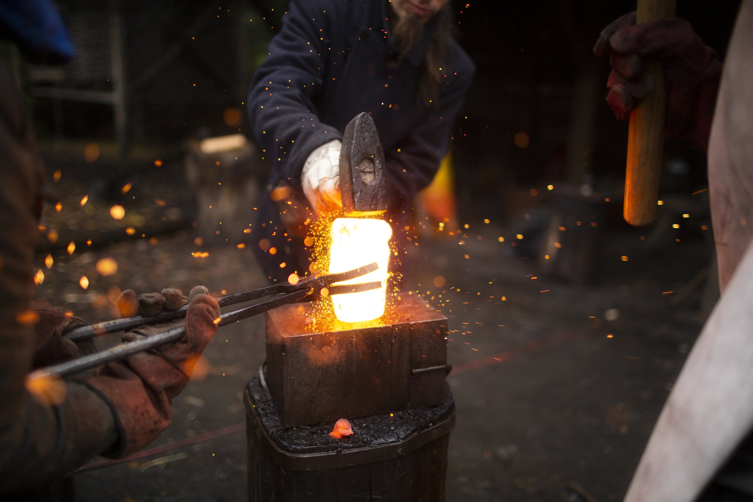 A blacksmith forges metal using a hot flame and sparks flying around, with a person holding tools on each side in a workshop.