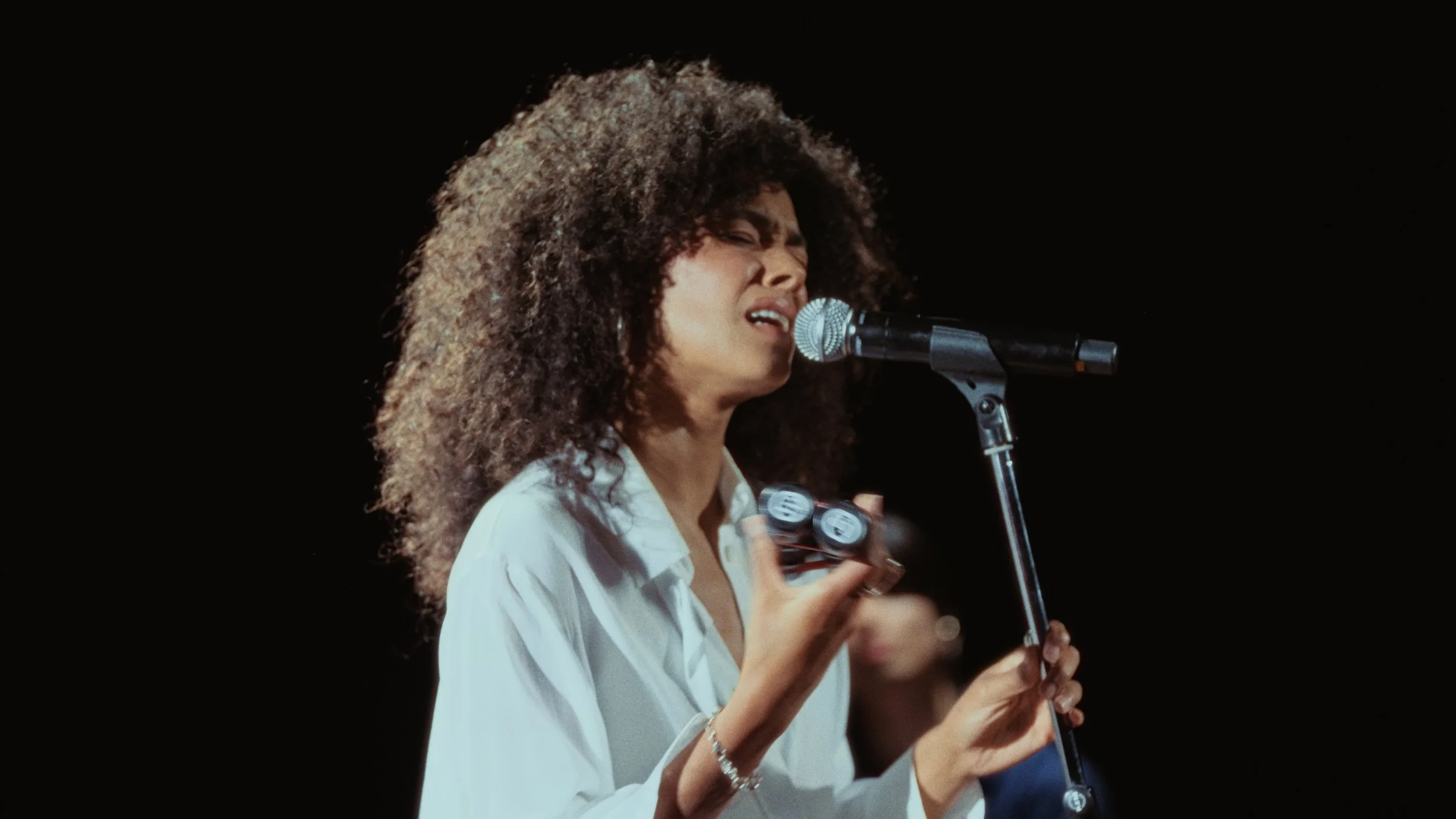 A woman with curly hair singing into a microphone on stage, holding a violin in her hand, wearing a white shirt.