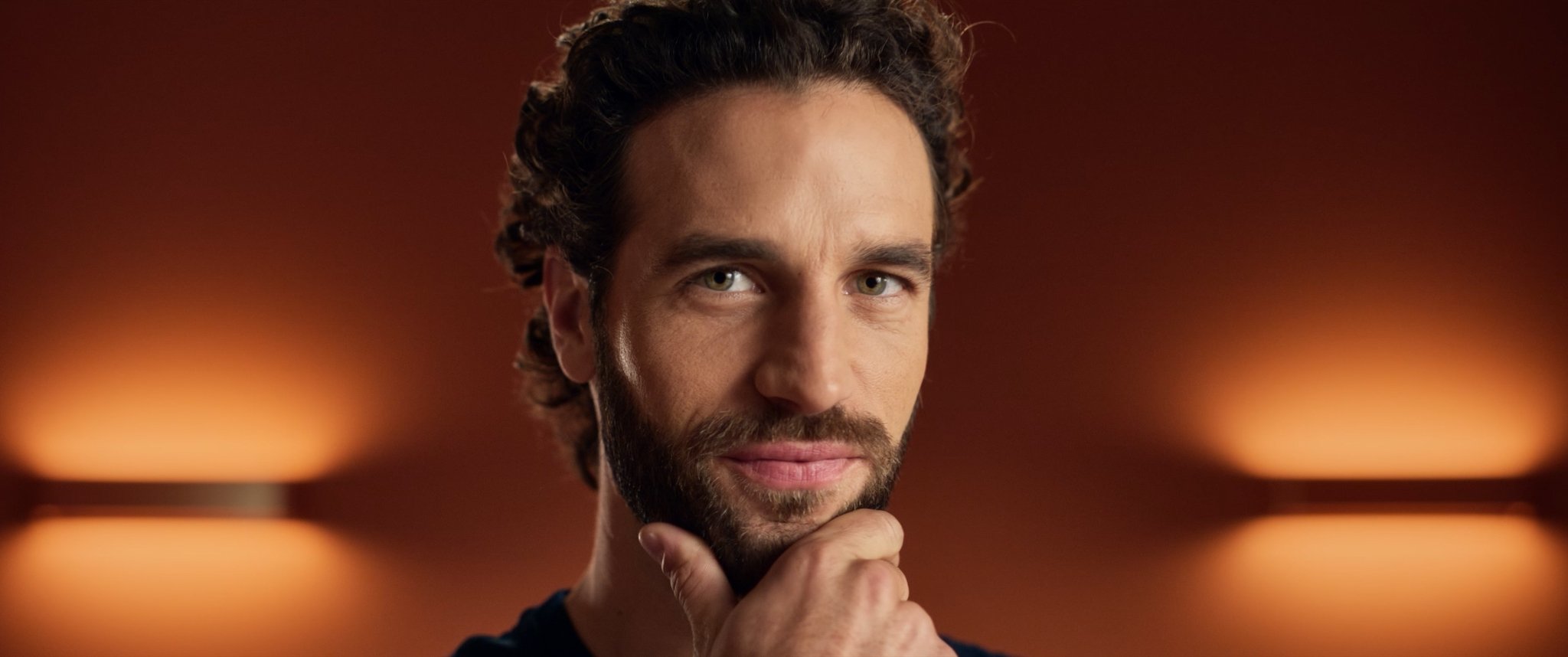 Portrait of a man with dark curly hair and beard, touching his chin thoughtfully, with warm blurred lighting in the background.