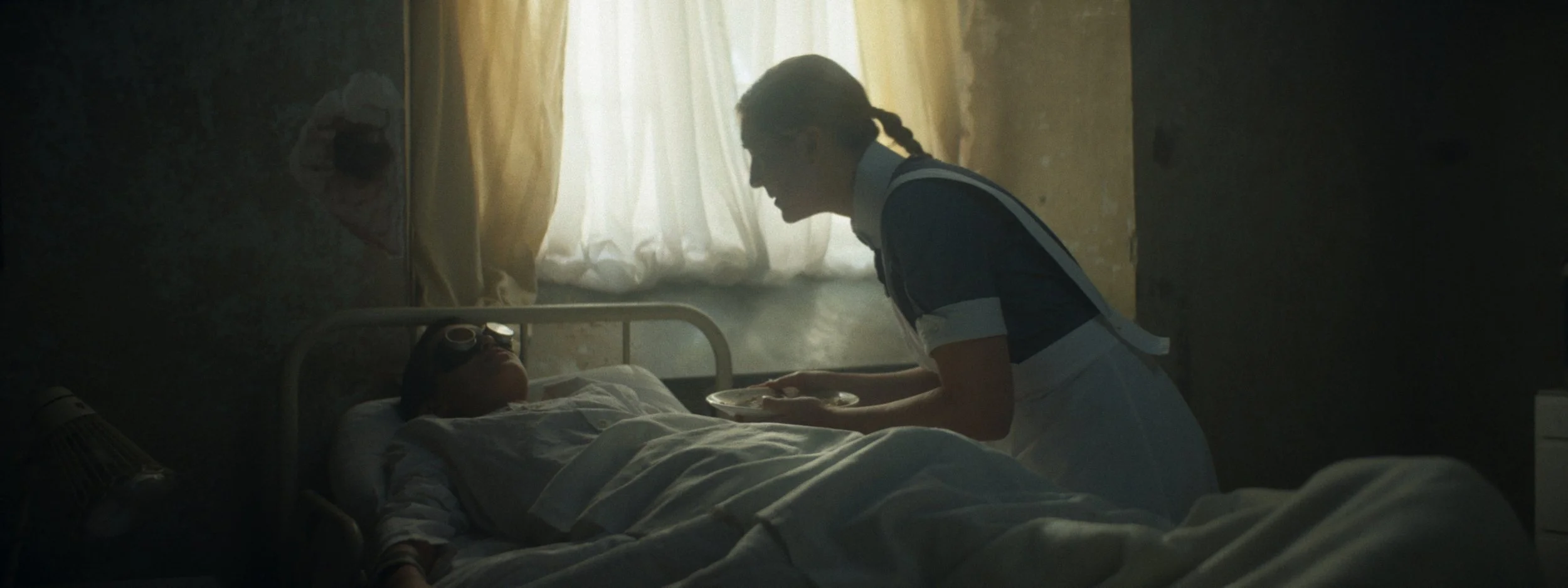 A nurse attending to a patient lying in bed in a dimly lit room with yellow curtains.