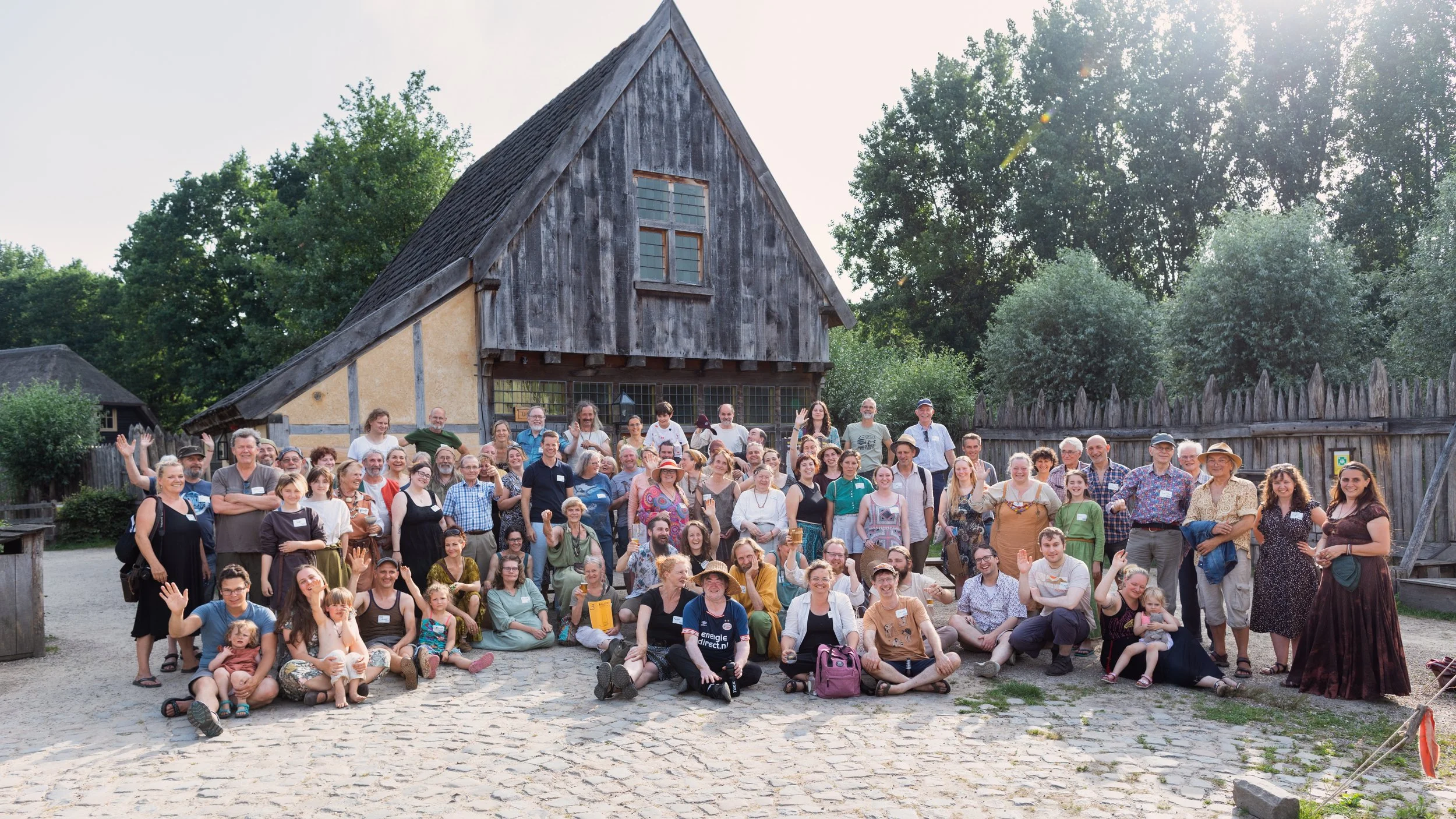 A large group of people gathered outdoors in front of a historic wooden building with a barn-like appearance, surrounded by trees."