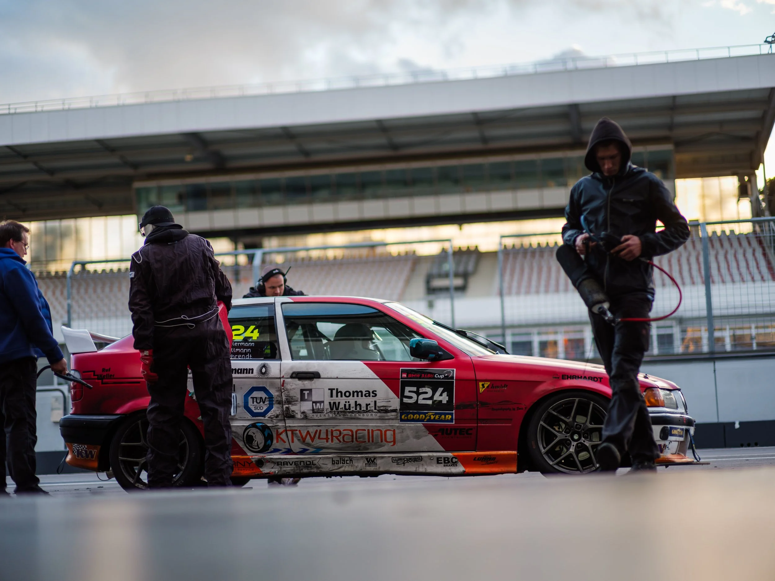 A race car team preparing a red and white race car at a racetrack, with team members around it and one person working on the tire, under an overpass with cloudy skies.