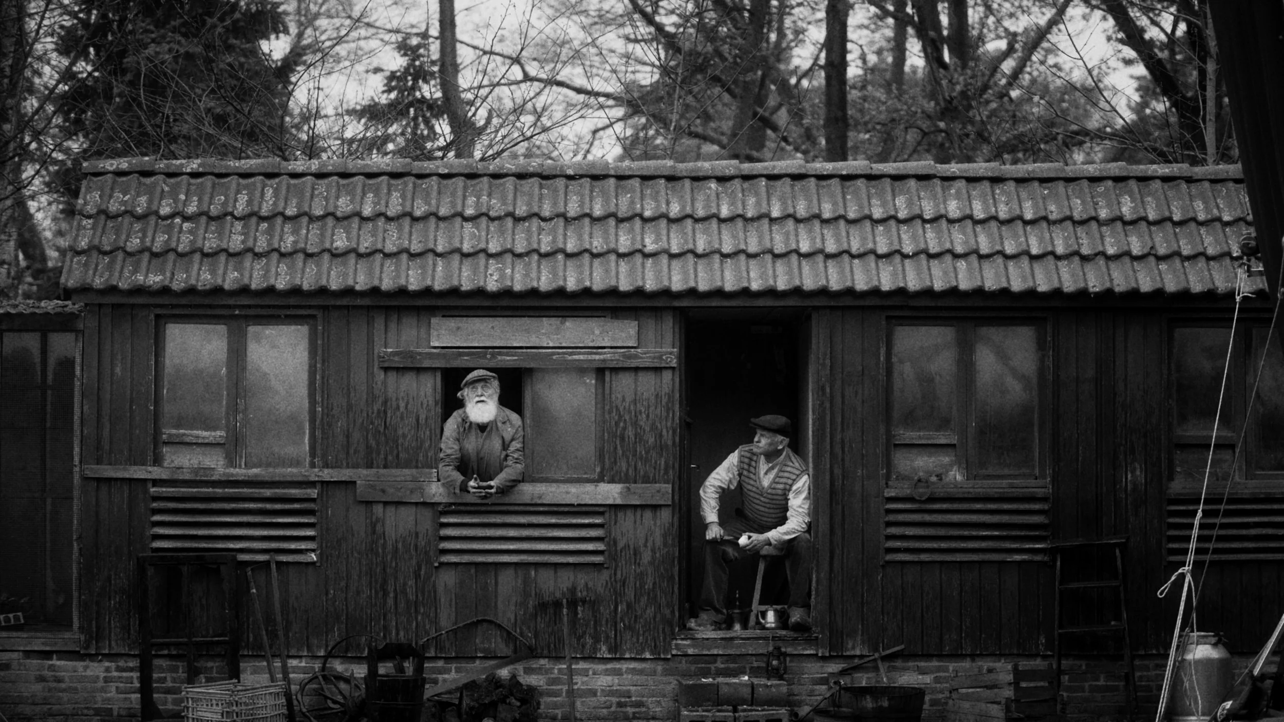 An old wooden house with a tiled roof and two elderly men standing on a balcony, one leaning against the railing and the other sitting on a chair. The house has windows with wooden shutters and a slatted lower section. Tall trees are visible in the b