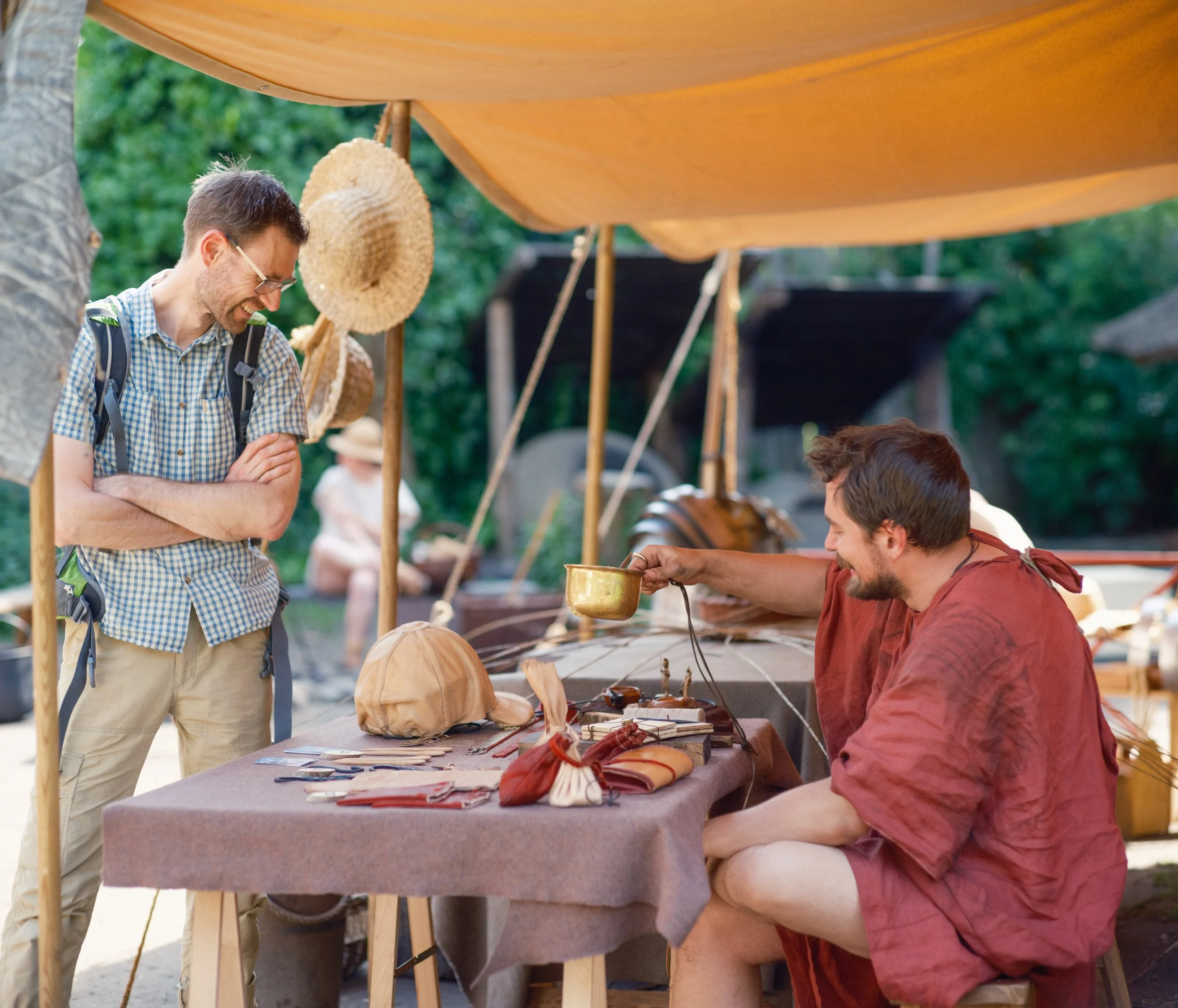 Two men at an outdoor craft market stall displaying handmade leather goods and accessories, with green trees in the background.