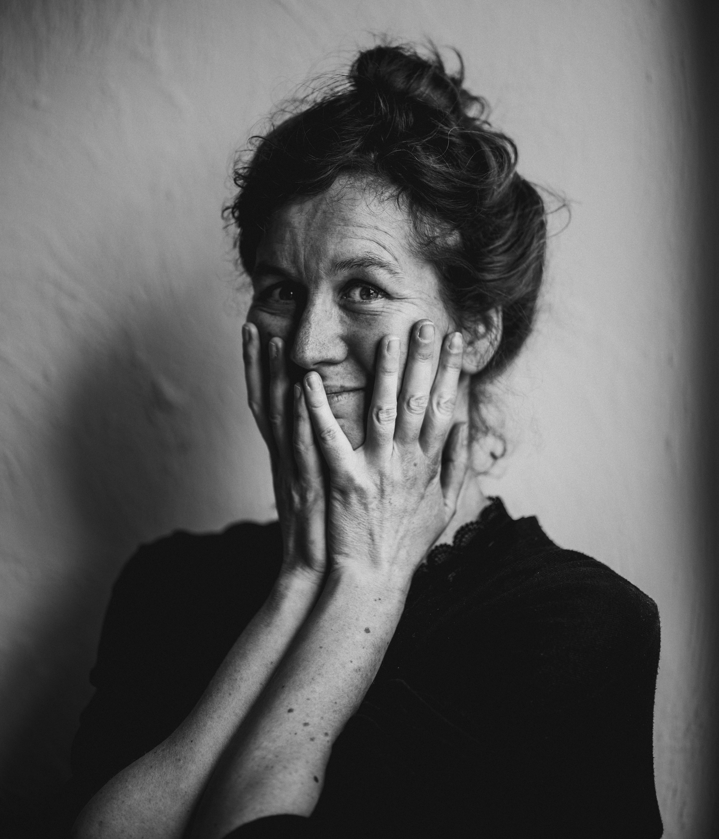A black and white close-up photo of a woman with curly hair, smiling and resting her face in her hands against a plain background.