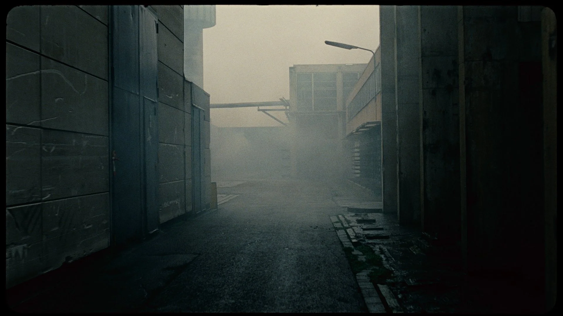 A foggy alleyway between industrial buildings with metallic walls on the left and dark walls on the right, a streetlight, and a distant building visible in the background.