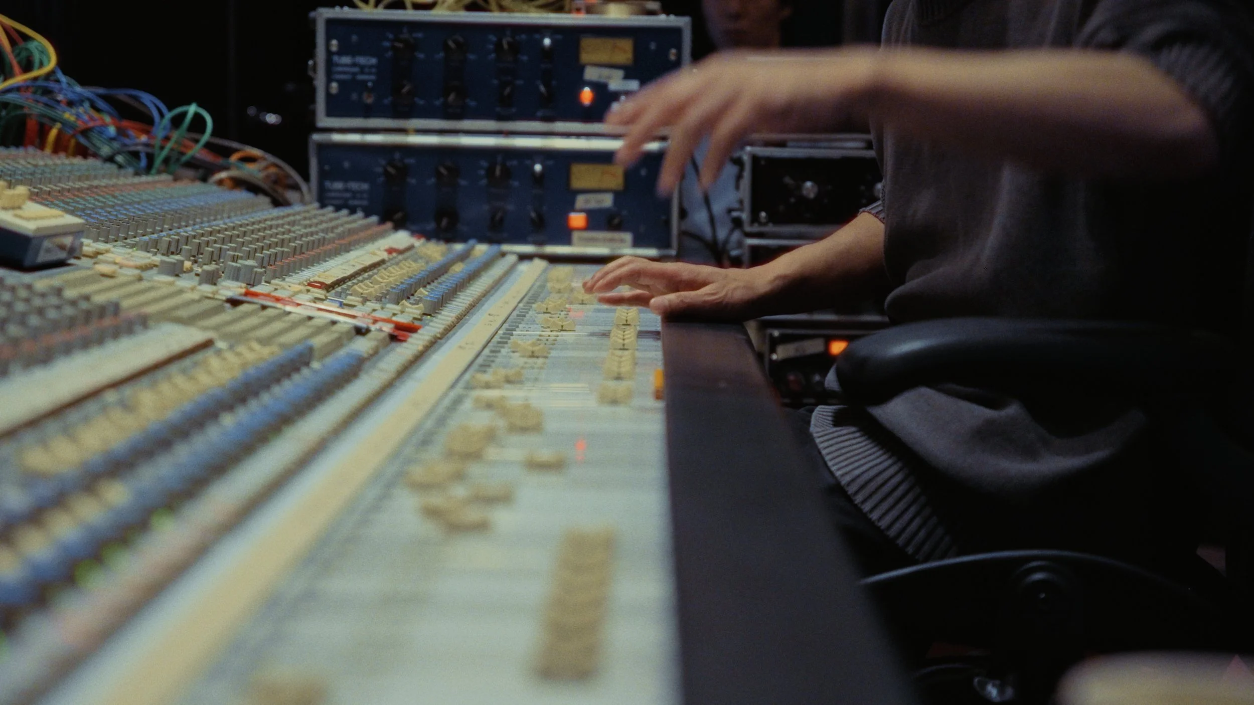 A person working on an audio mixing console with numerous knobs and sliders, alongside electronic equipment and cables.