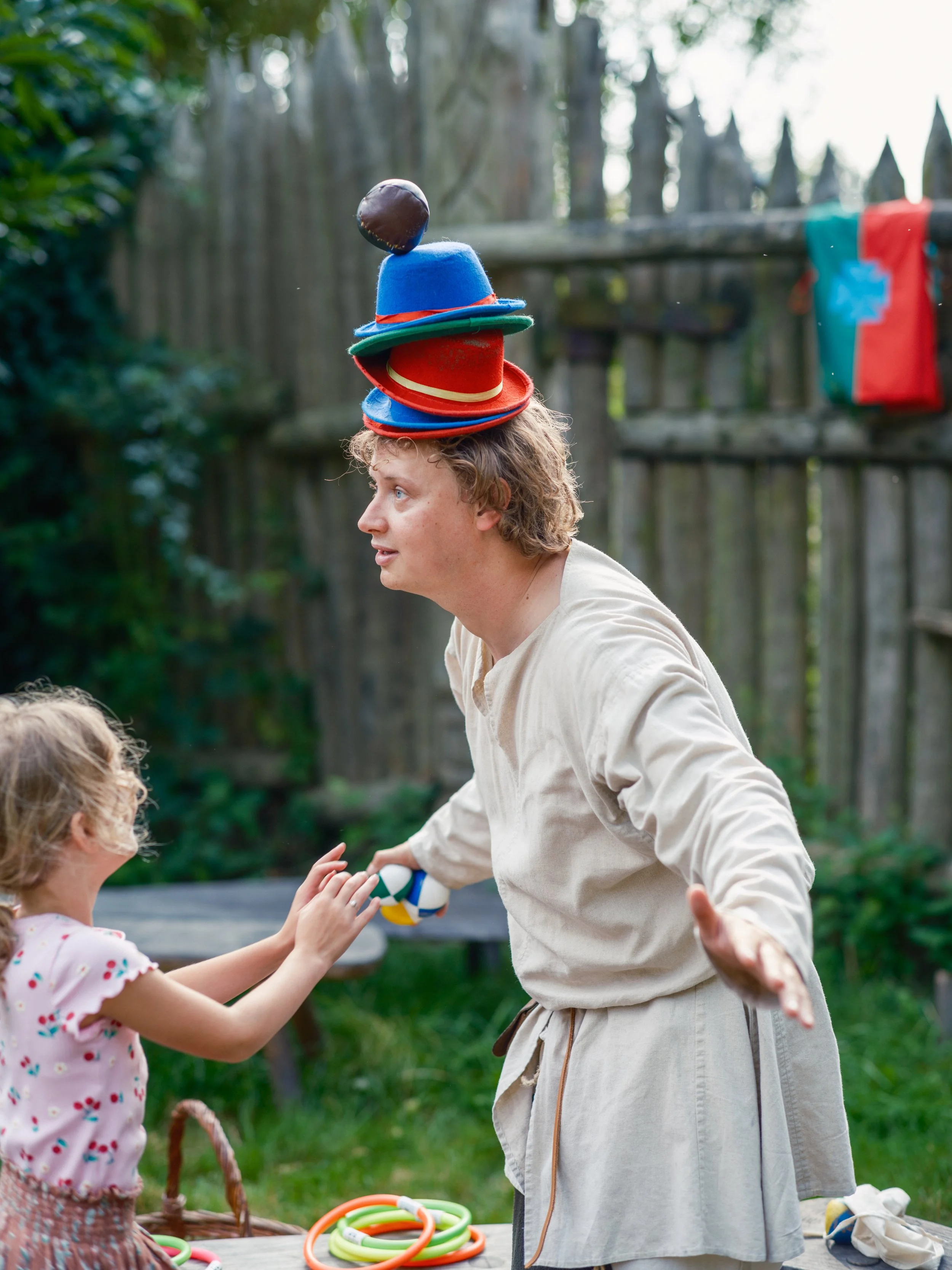 A woman and a young girl playing with a colorful ball outside. The woman is balancing several hats on her head and holding a ball out to the girl. There is a table with colorful plastic hoops and other items, and a wooden fence in the background.