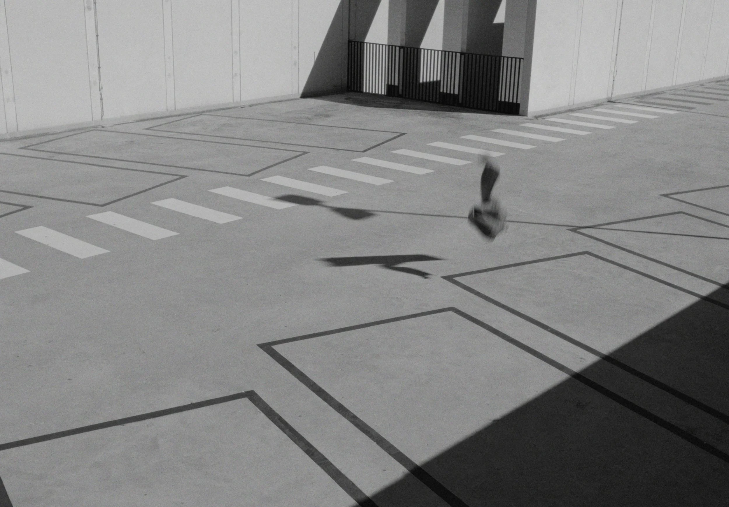 Empty gray parking lot with white parking space lines, a small black fence, a concrete wall, and shadows cast by nearby structures.