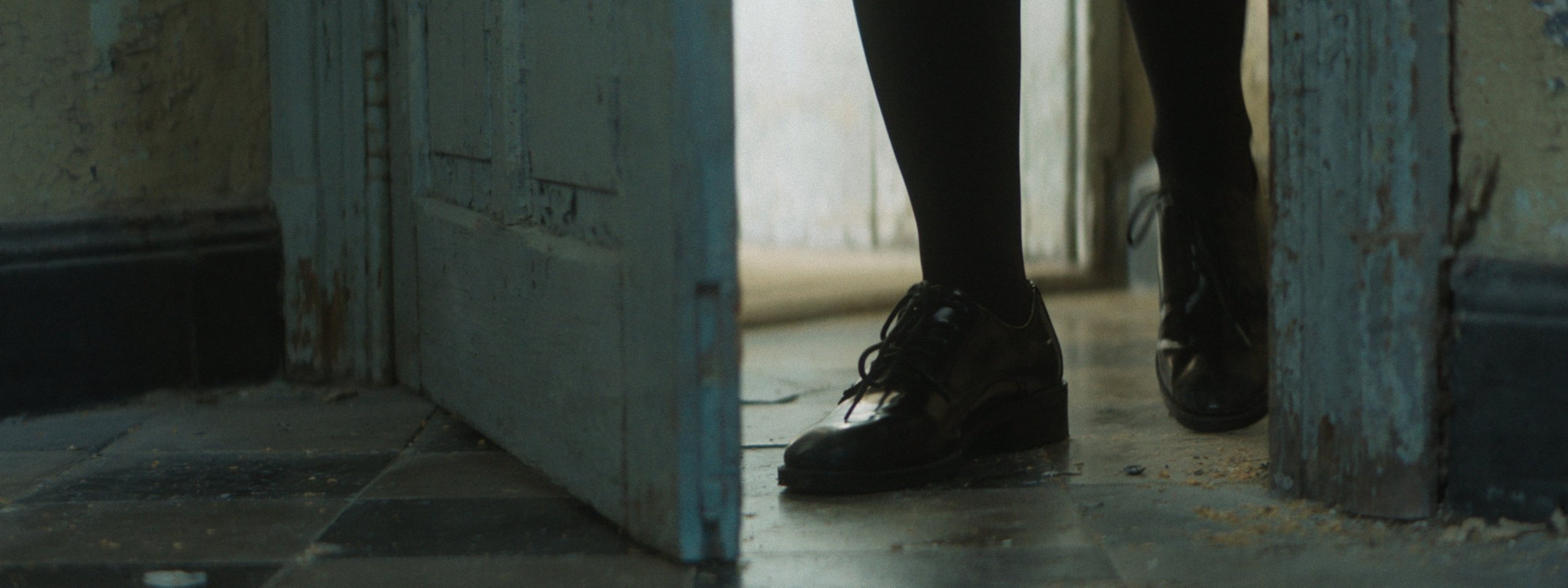 A person wearing black clothes and black shoes walking through a slightly open door in a dimly lit room.