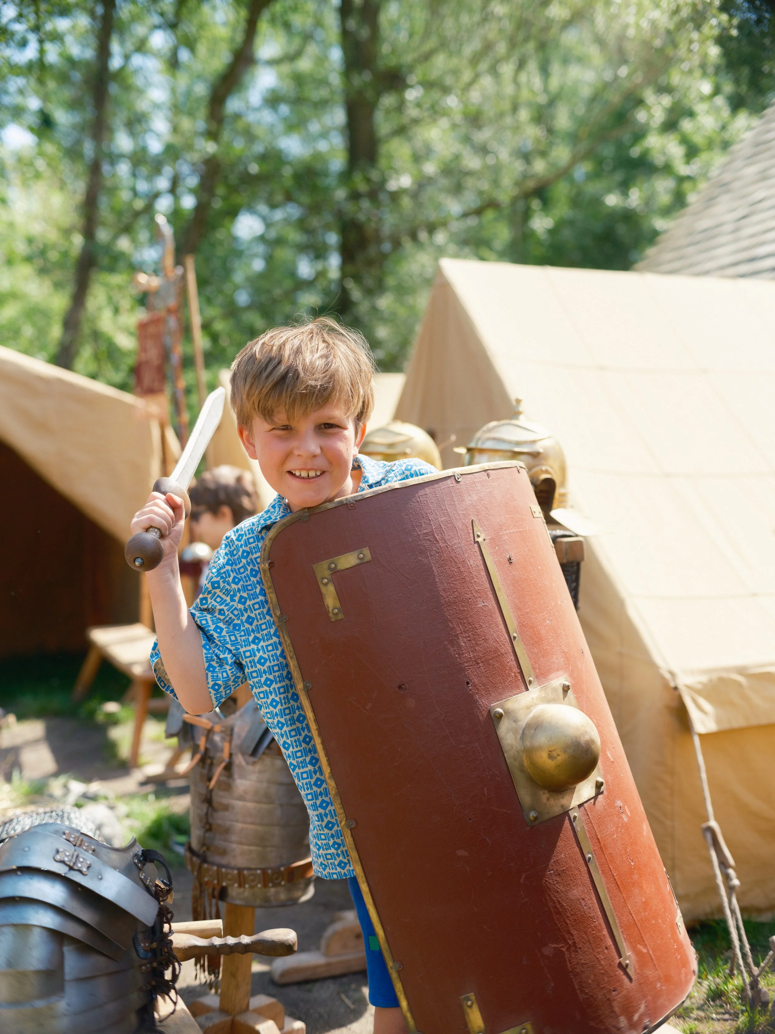 A young boy wearing a blue shirt with a pattern, holding a large wooden shield with brass fittings, standing outdoors at a medieval reenactment or fair, with tents and green trees in the background.
