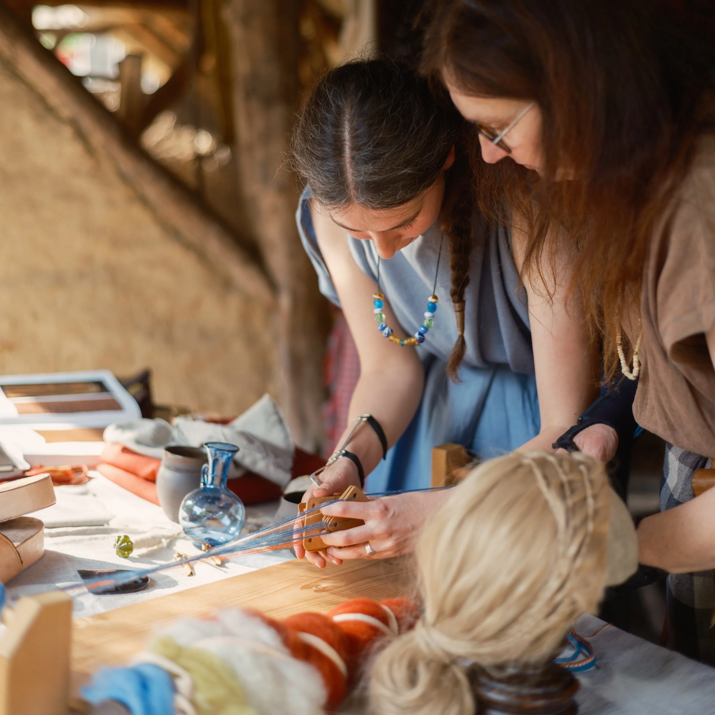 Two women with long brown hair and glasses working together on a craft project at a table, with yarn, a glass globe, and wooden objects in a cozy indoor setting.