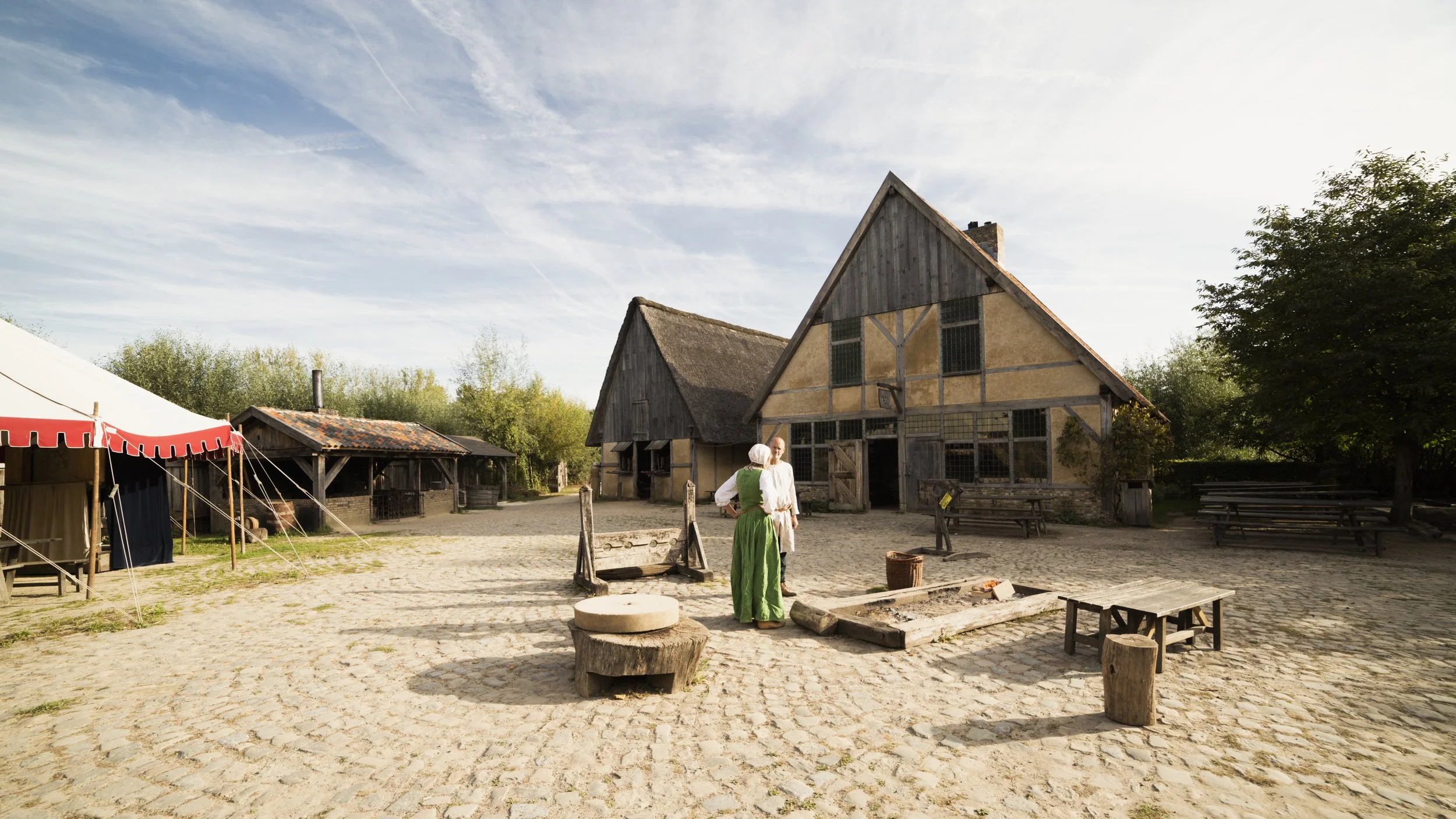 Two women dressed in historical clothing conversing in a medieval village setting with wooden buildings, tents, and cobblestone ground.