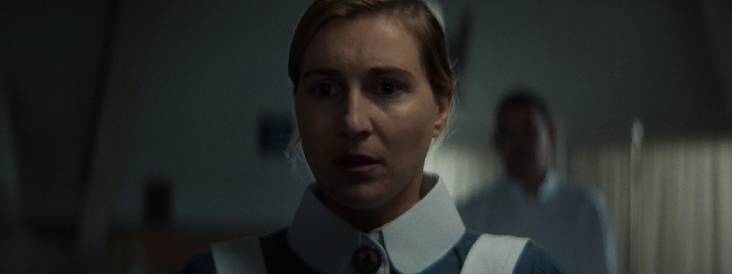 Close-up of a woman in a nurse's uniform looking concerned, with a person blurred in the background.