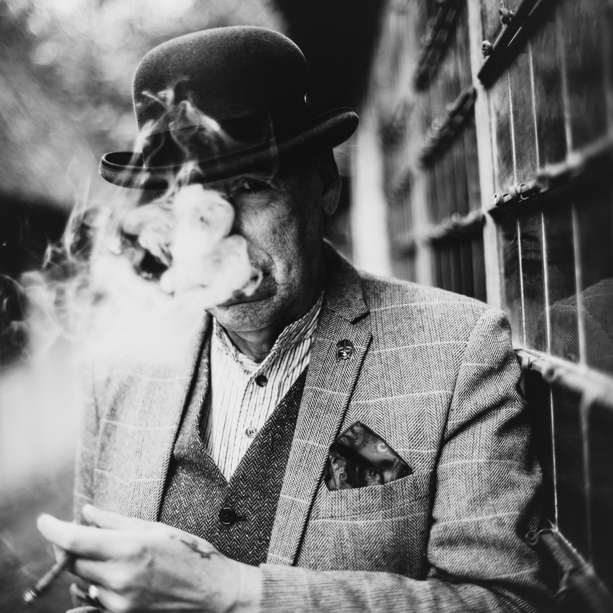 A person wearing a hat smokes a cigar, with smoke visible in the foreground, against a blurry background.