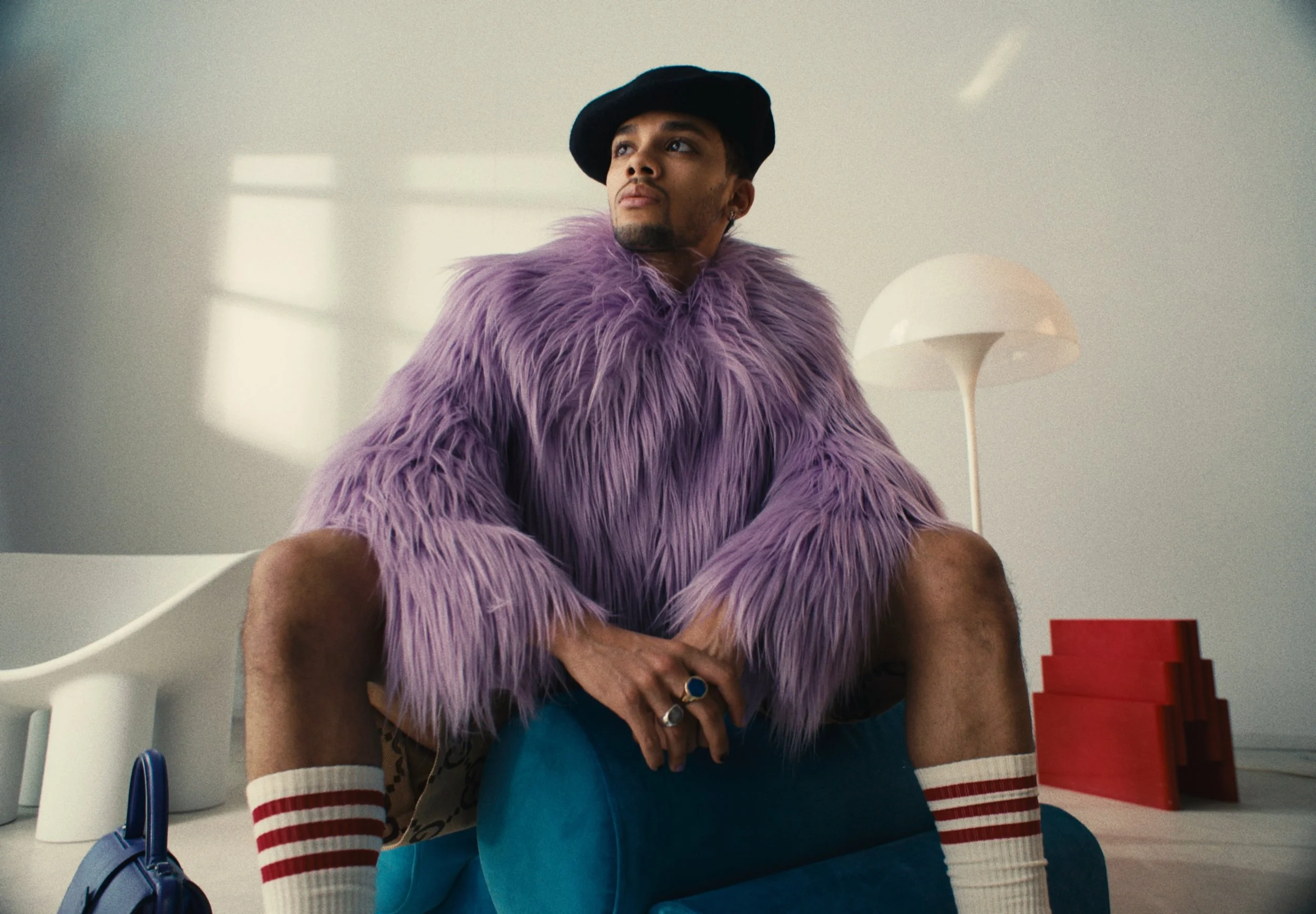 Person wearing a purple furry jacket, a black beret, and knee-high socks with red stripes, sitting on a table in a modern, minimalistic room with white walls and a white lamp.