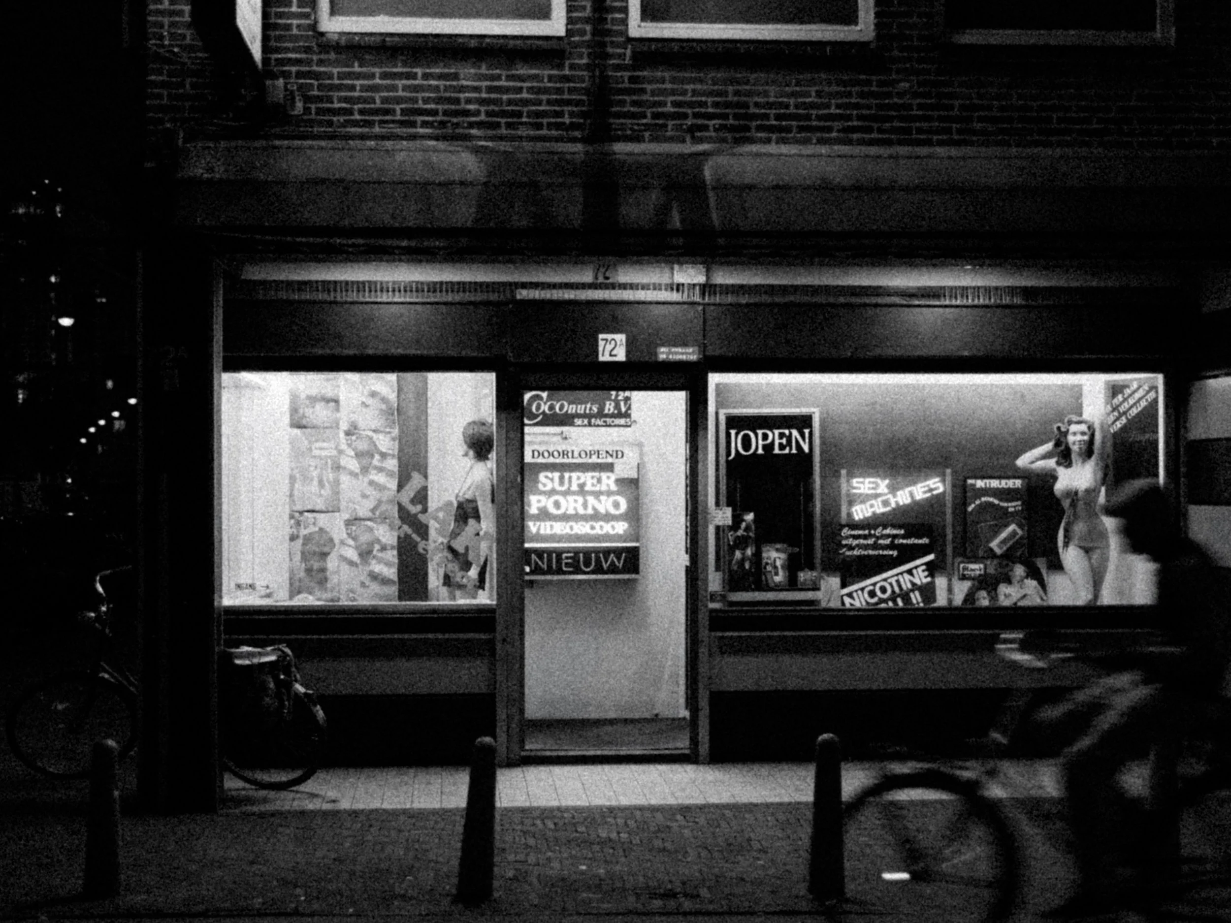 A storefront with signs advertising adult content and cigarettes, visible through a glass window and door, with a person standing outside and a bicycle parked nearby at night.