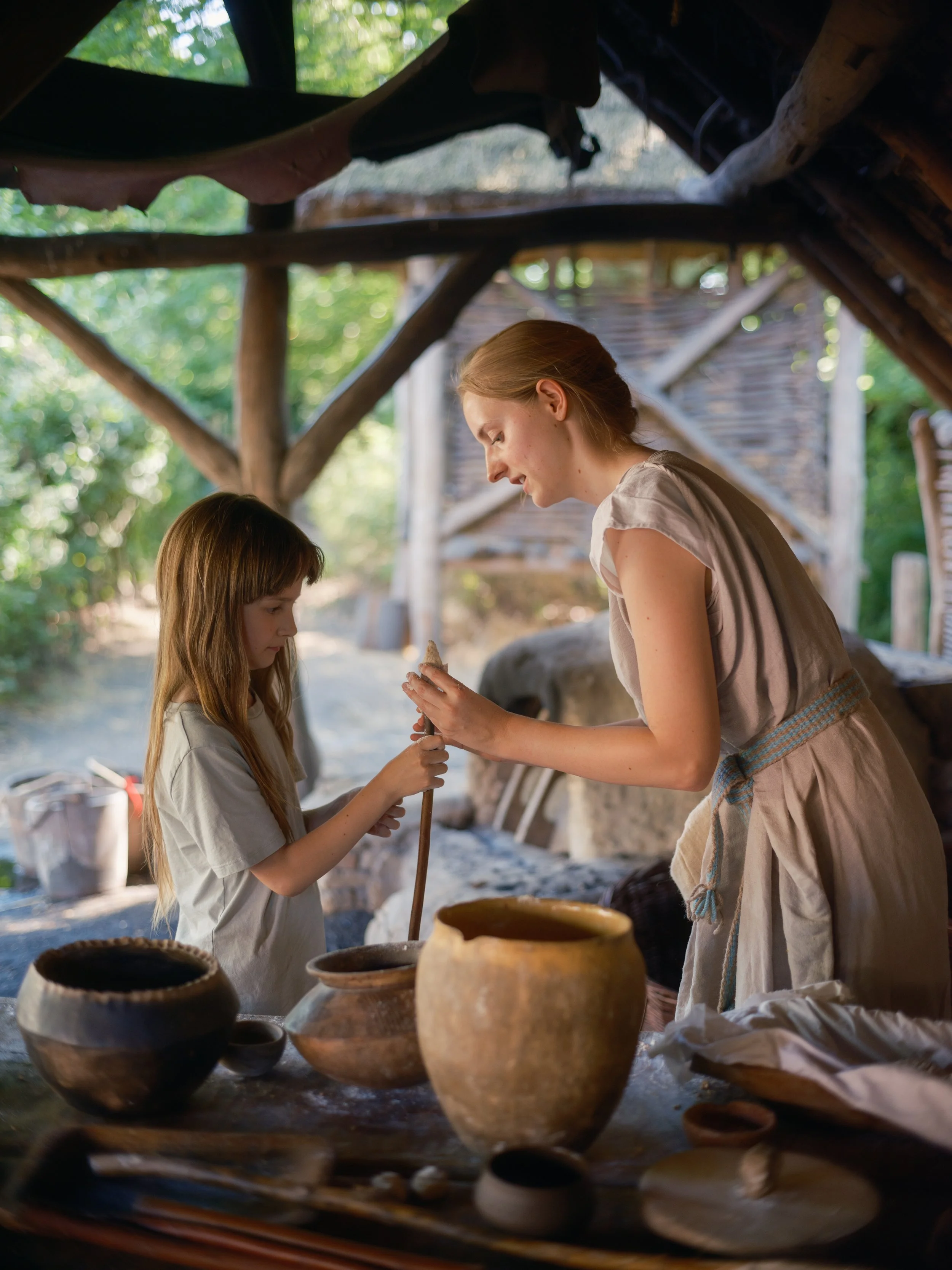 A woman and a young girl in historical clothing at an outdoor pottery stall, surrounded by clay pots and jars on a wooden table, under a rustic shelter with green trees in the background.