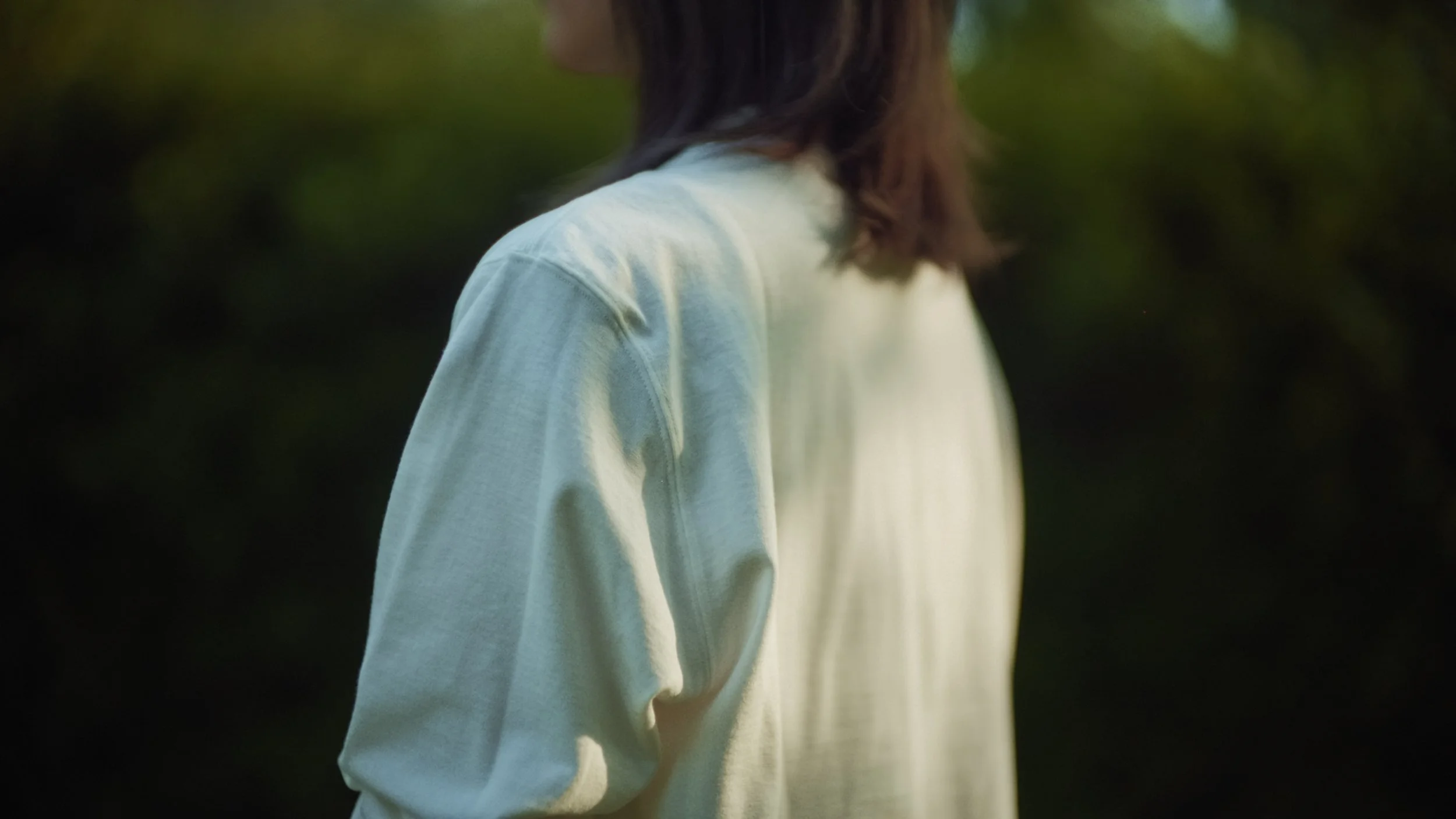 Close-up of a person with dark brown hair wearing a white long-sleeve shirt, standing outdoors with greenery in the background.