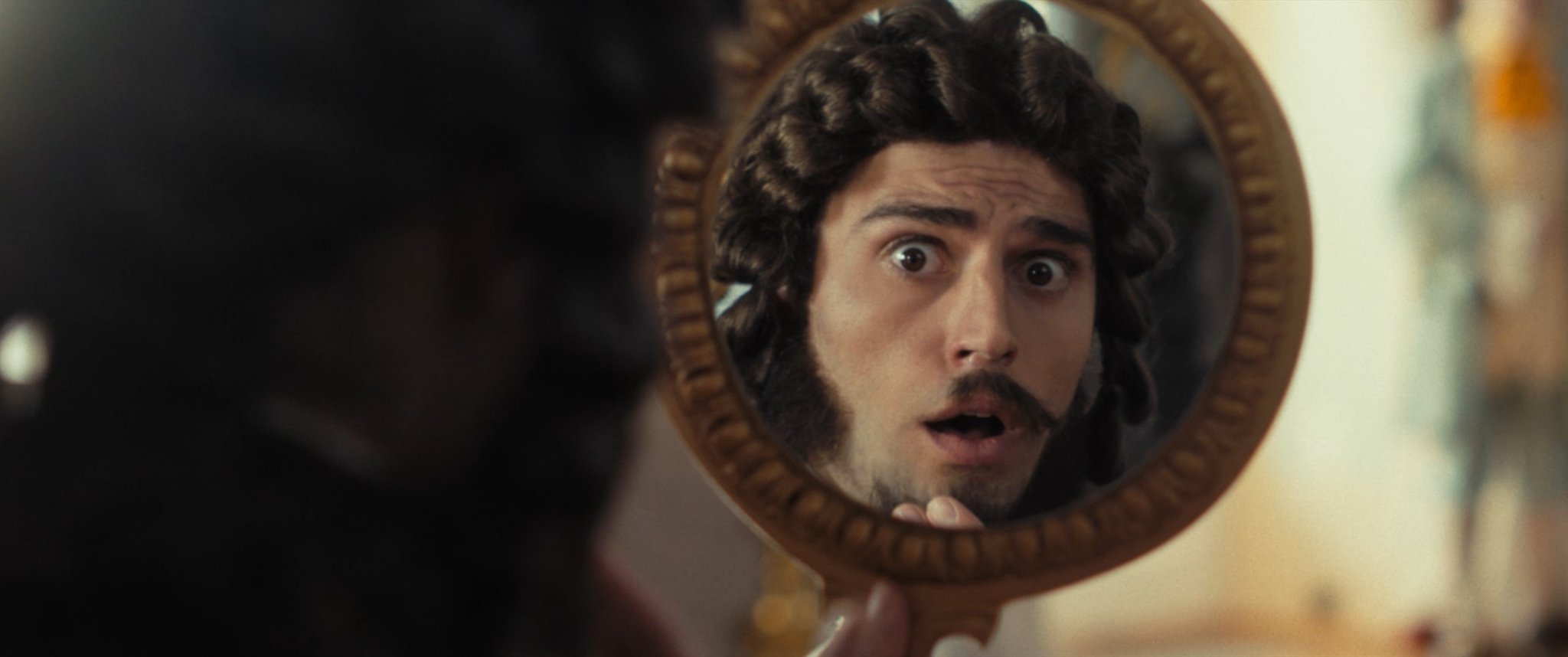 A man with curly dark hair and sideburns looking surprised at his reflection in a handheld mirror.