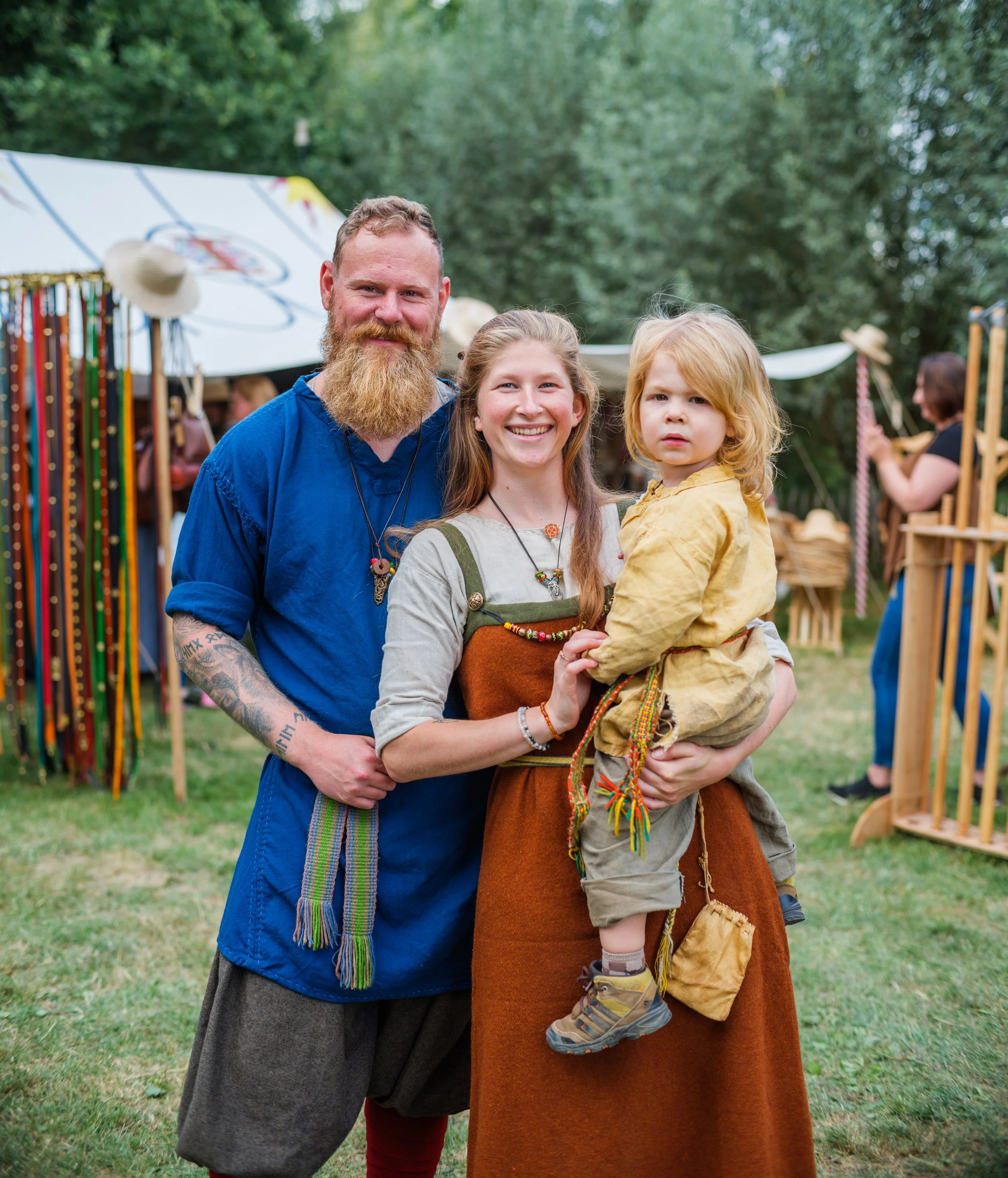 A family of three standing outdoors at a festival or fair, with various booths and trees in the background. The man has a red beard, wearing a blue shirt, and the woman has long hair, wearing a brown dress, holding a young boy with curly blonde hair.
