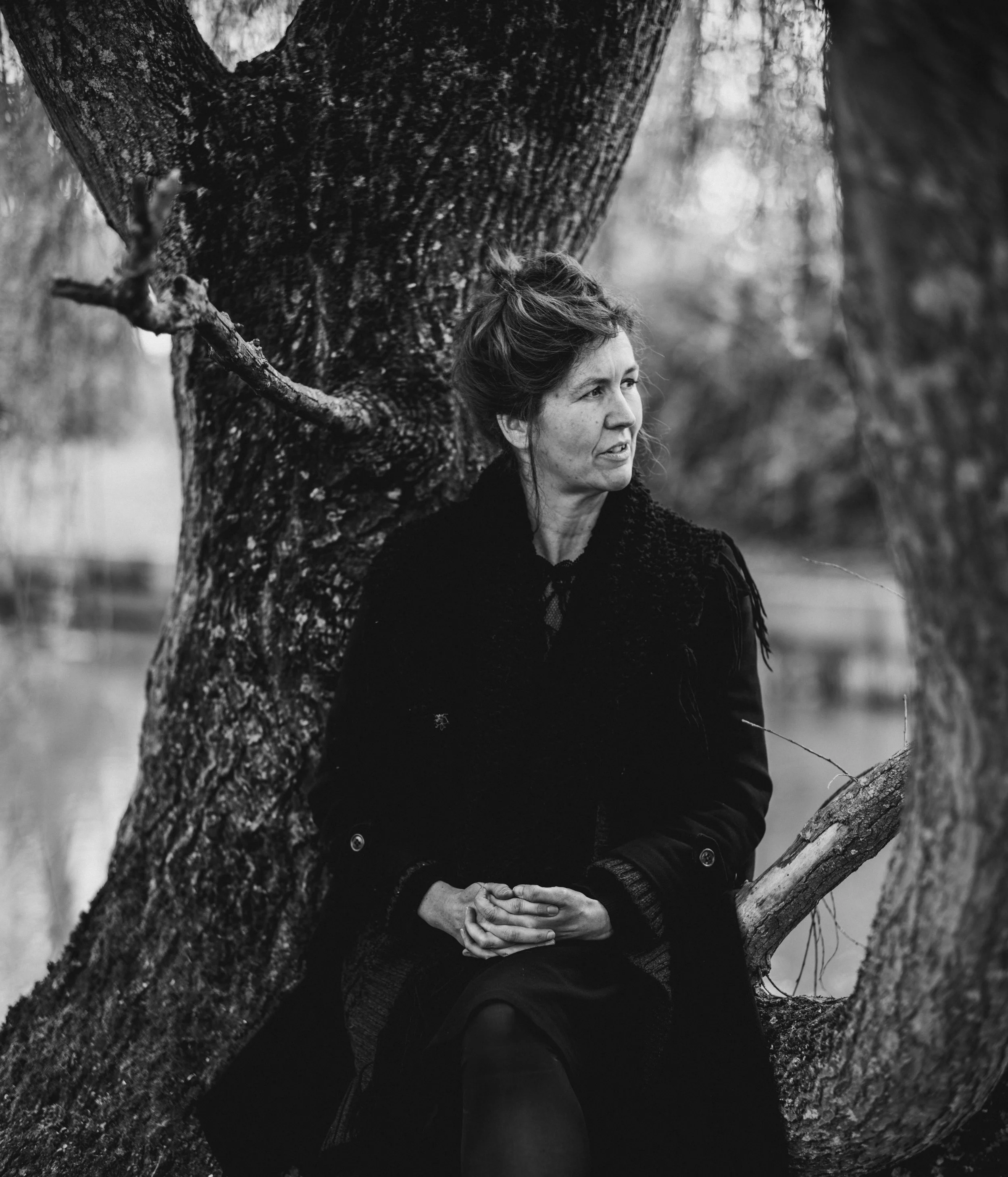 Black and white photo of a woman sitting on a large tree trunk in an outdoor park or forest setting, with a serene expression and hands clasped in her lap, surrounded by blurred trees and nature.