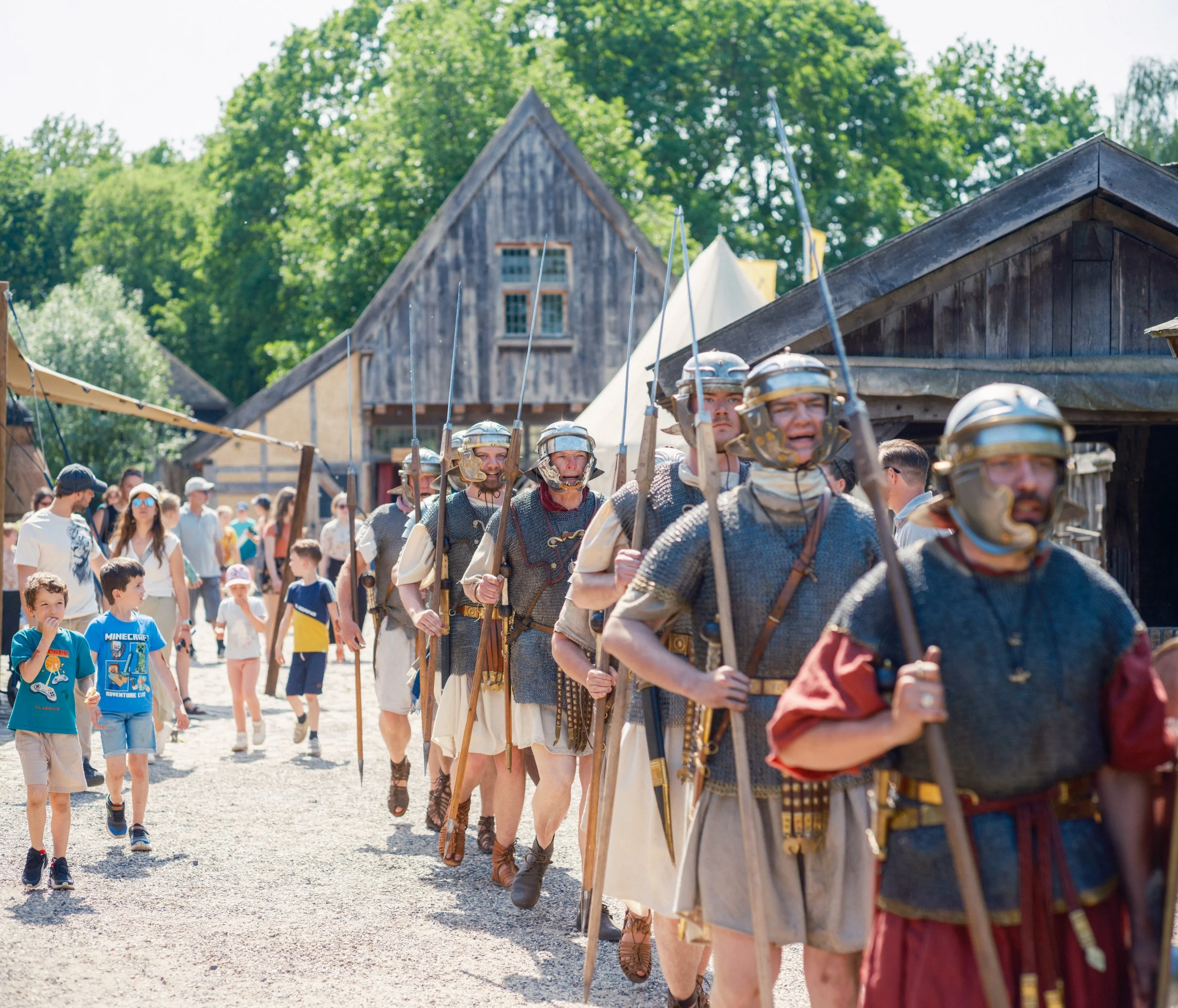 People dressed as Roman soldiers marching outdoors at a historical reenactment event with historical buildings and green trees in the background.