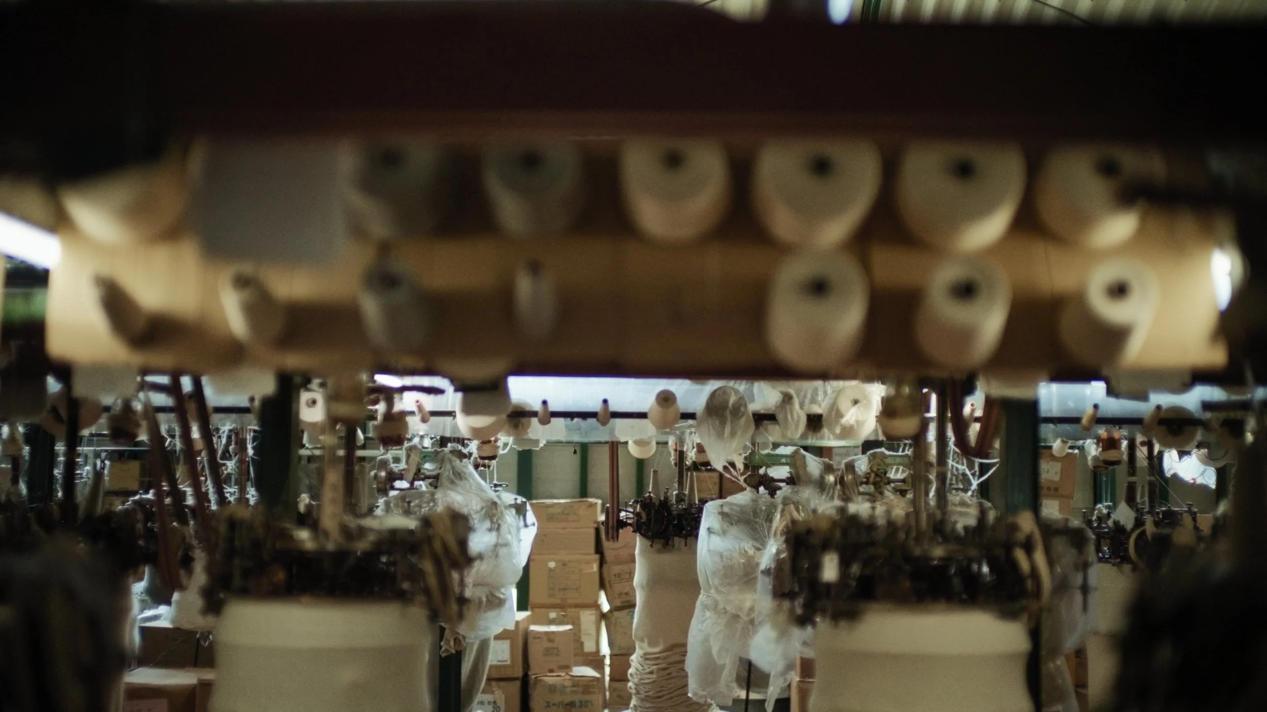 Close-up view of yarn spools and weaving equipment in a textile workshop, with shelves filled with boxes and materials in the background.