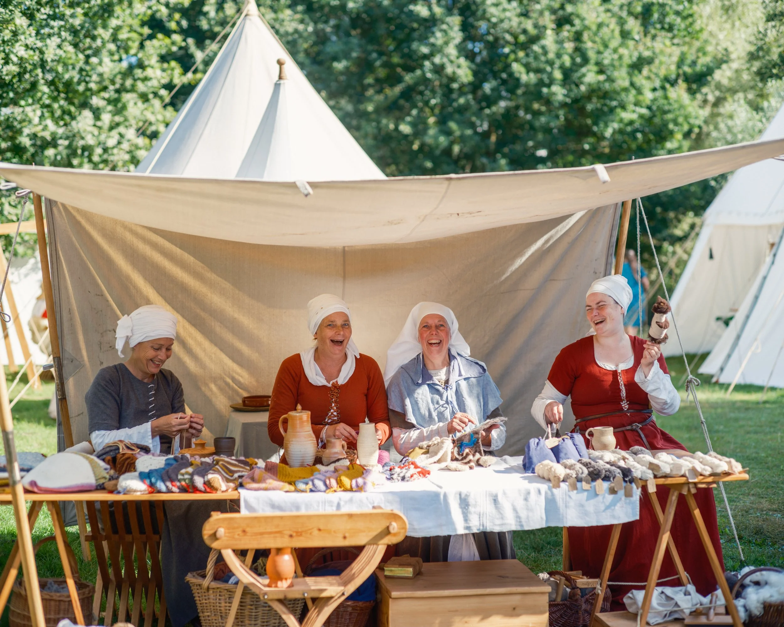 Women in traditional clothing sitting behind a table of handmade crafts at an outdoor market under a canopy in a park with trees.