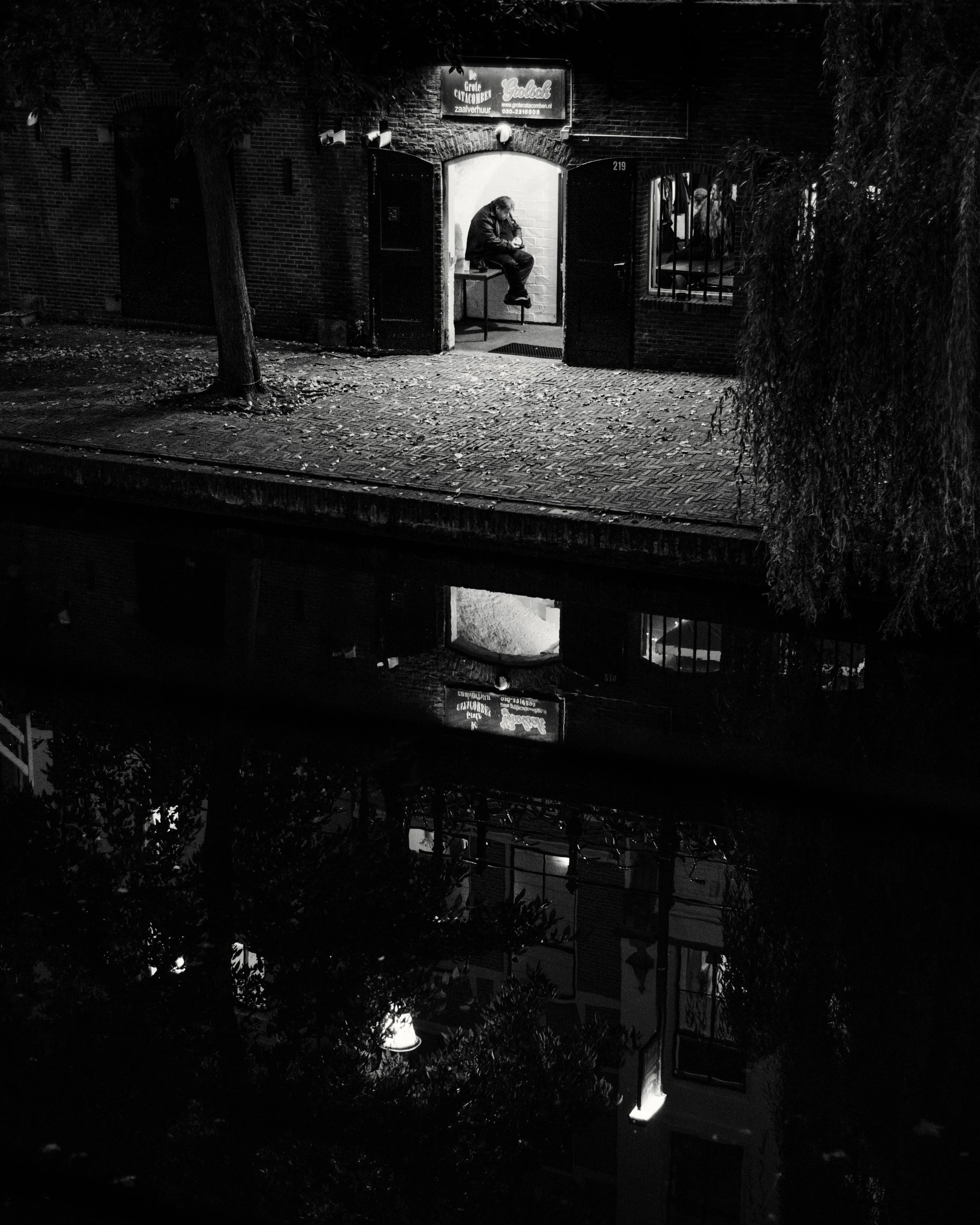 Nighttime scene of a man sitting inside a small brick building, illuminated by lights, with a large tree and a window with bars on either side. The image appears upside down.