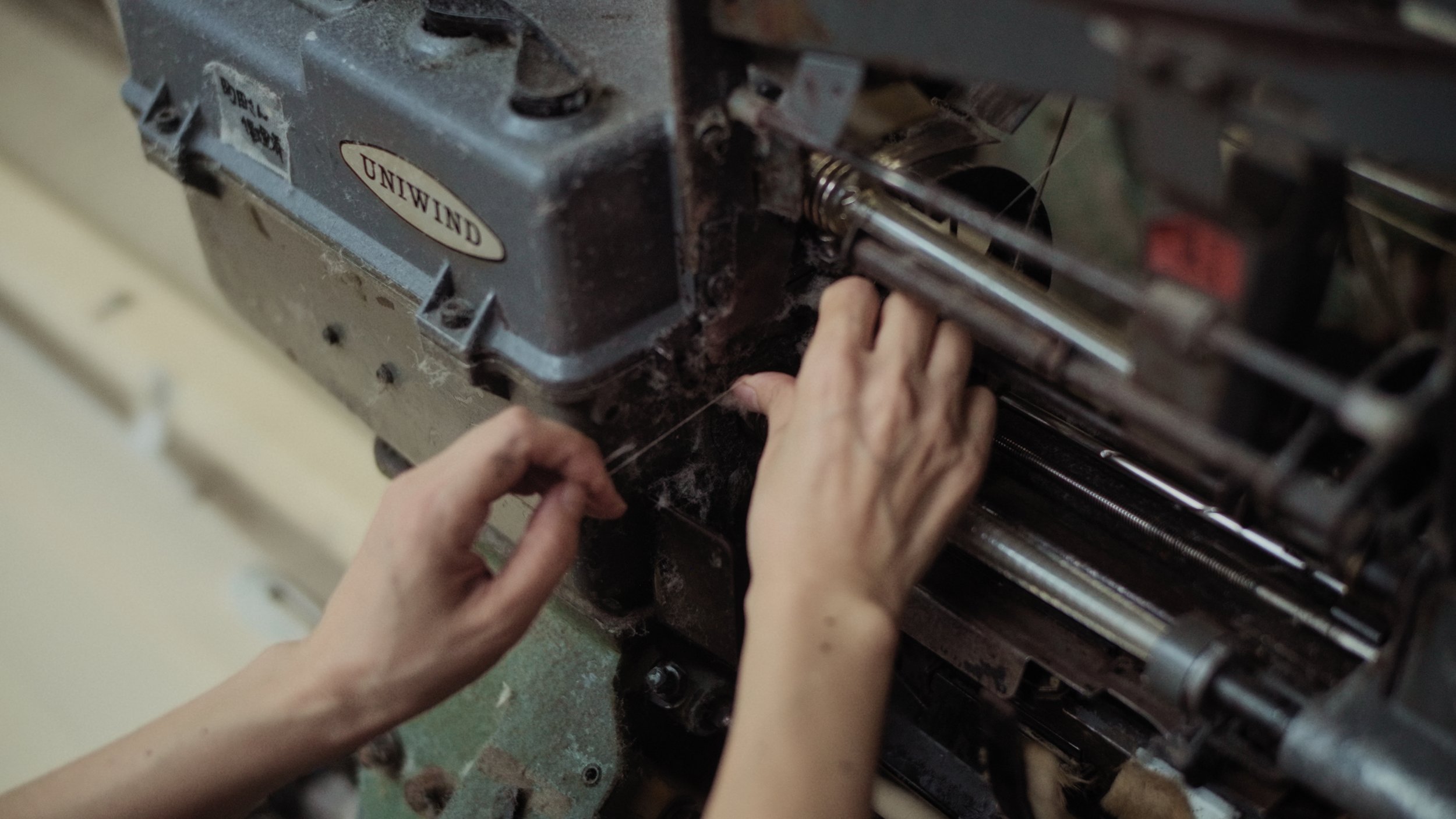 Hands working on a mechanical machine, possibly a sewing machine or industrial equipment, with a label that reads 'UNIWIND'.