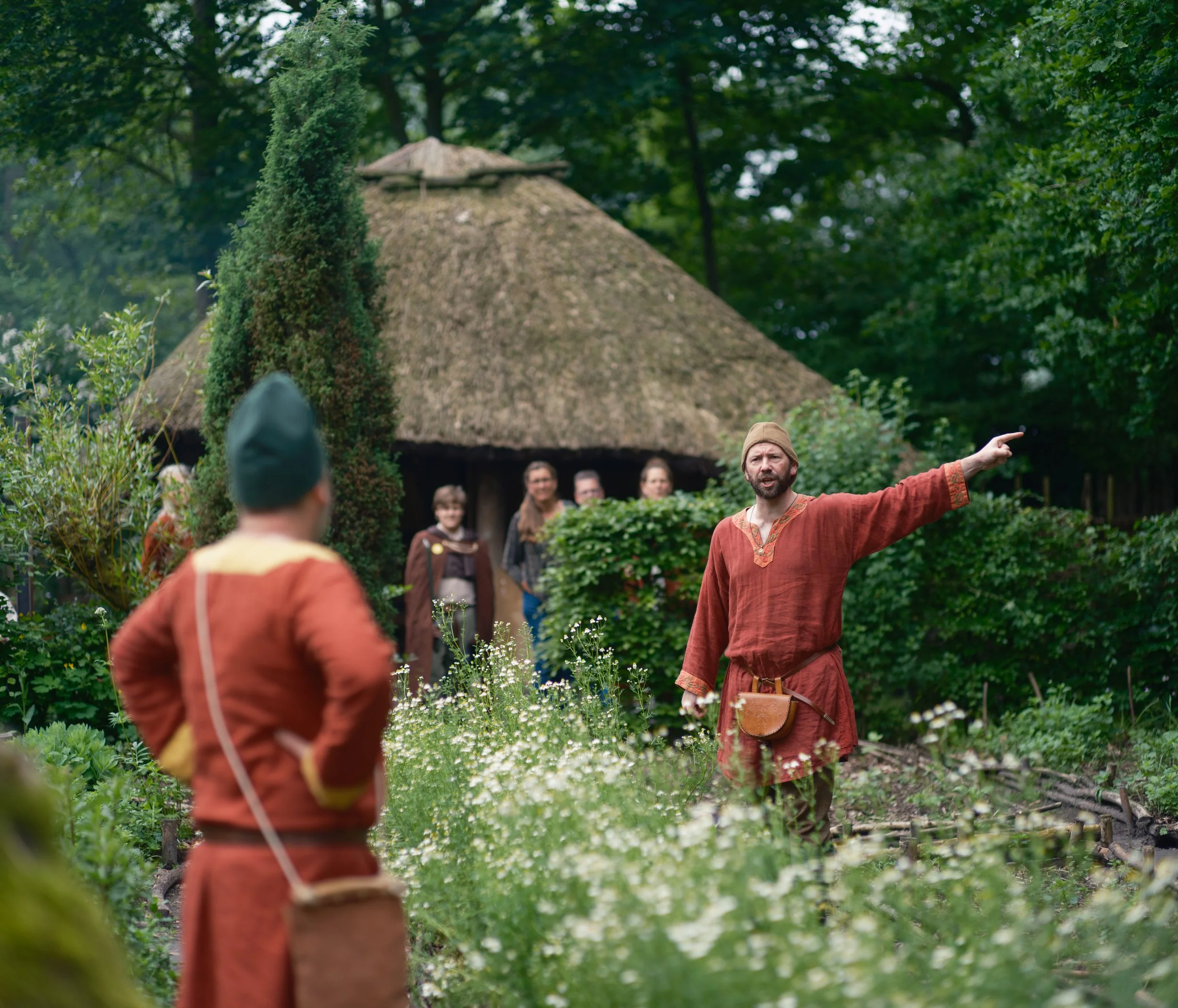 A person dressed in historical clothing giving a speech or performance outdoors, with a group of people watching in front of a thatched-roof structure surrounded by green trees and plants.