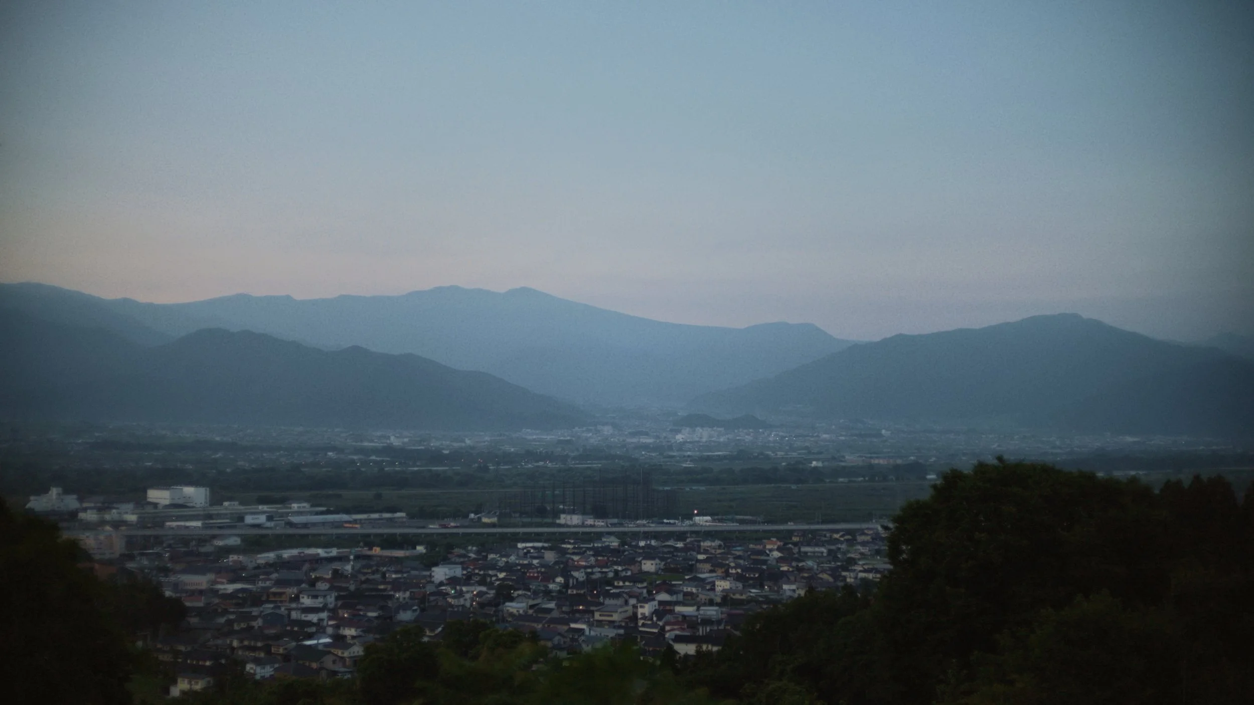 A cityscape with a residential neighborhood in the foreground, a highway, and a mountain range in the background under a cloudy sky.