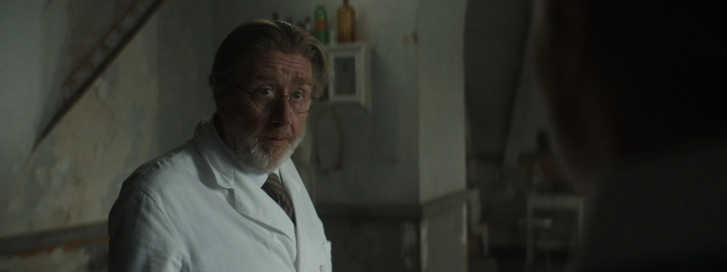 A middle-aged man with gray hair, glasses, and a beard, wearing a white lab coat, looking over his shoulder in a dimly lit room that appears to be a medical or laboratory setting.