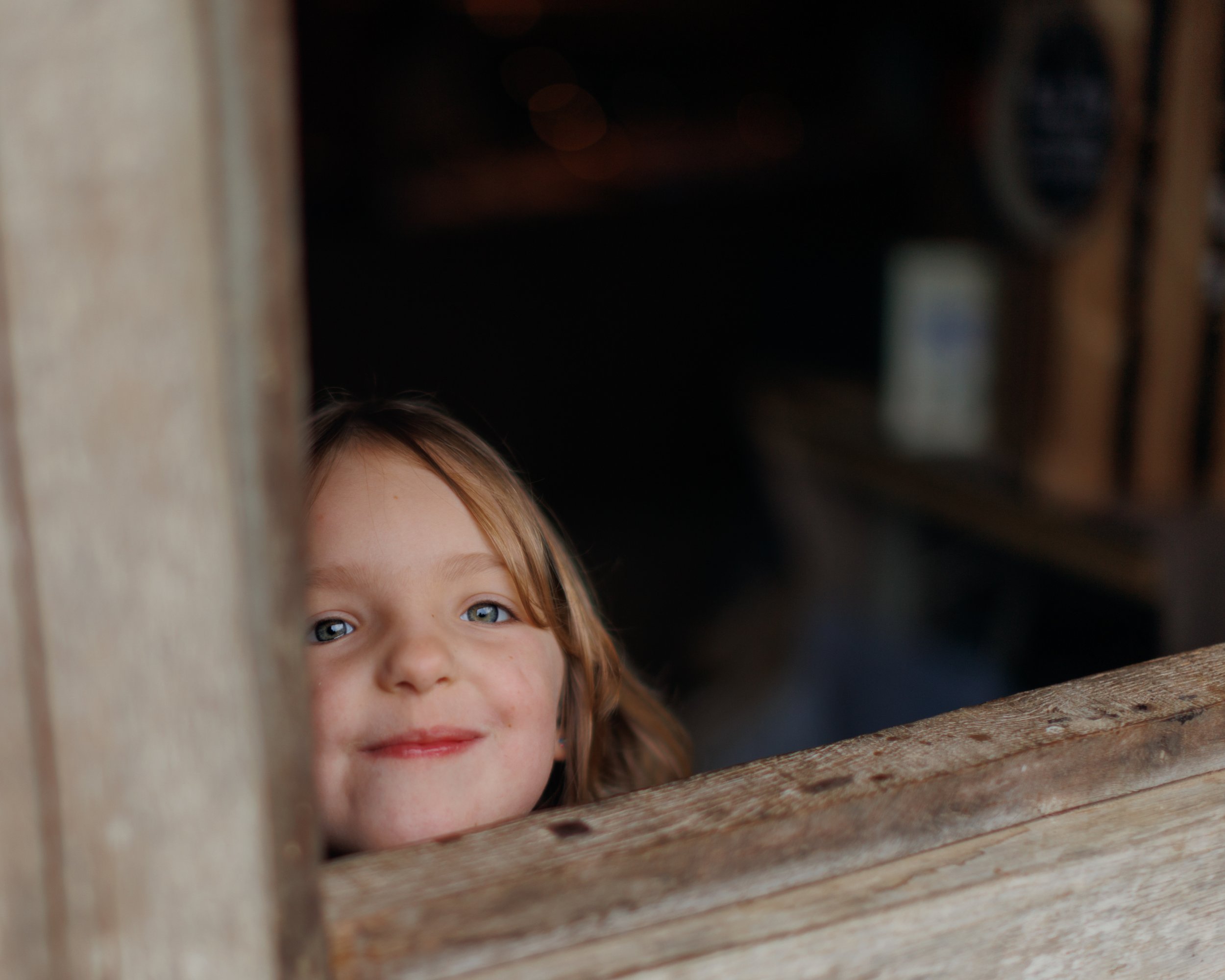 A young girl with blue eyes and red hair looking through a wooden fence or slats.