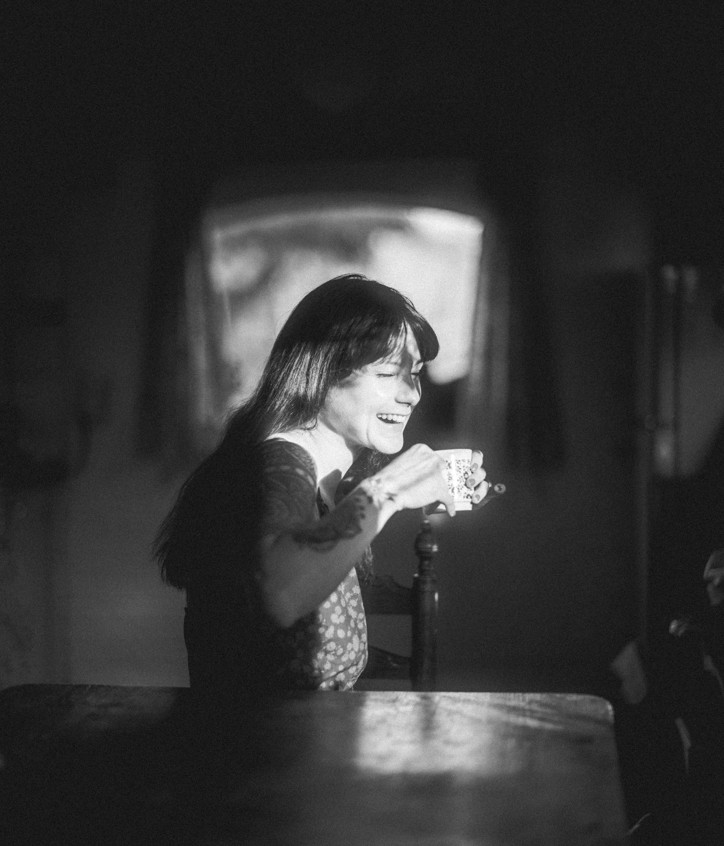 Black and white photo of a woman with long hair smiling and drinking from a cup, sitting at a table in a dimly lit room with an arched doorway in the background.