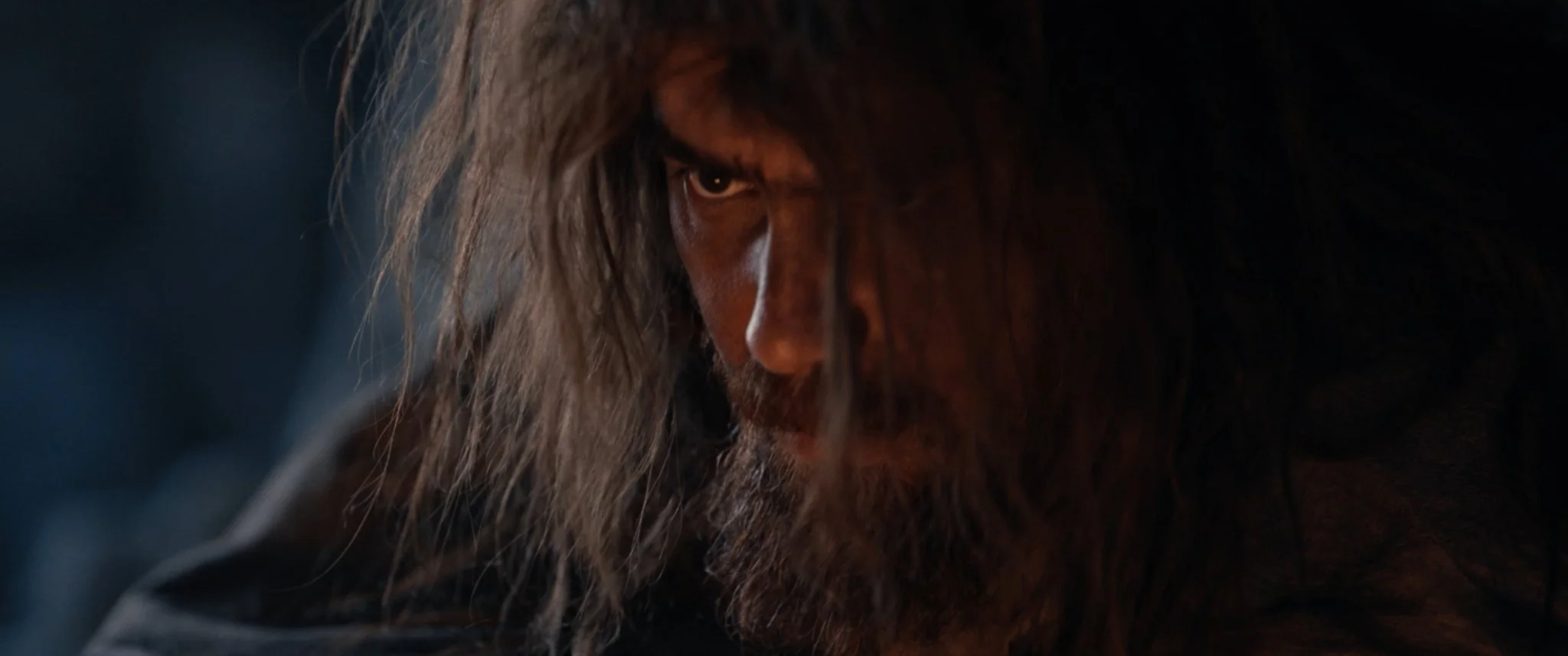 Close-up of a man with long hair and a beard, looking intensely to the side with a dark background.