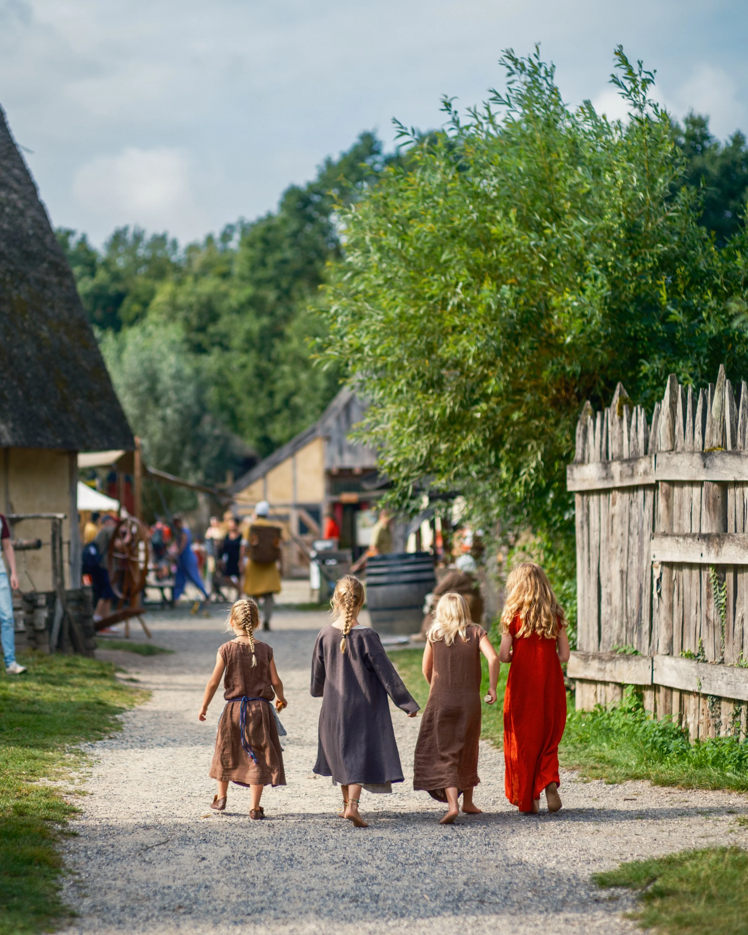 Five children dressed in medieval-style clothing walking down a dirt path in a historic village with wooden fences and thatched-roof buildings.
