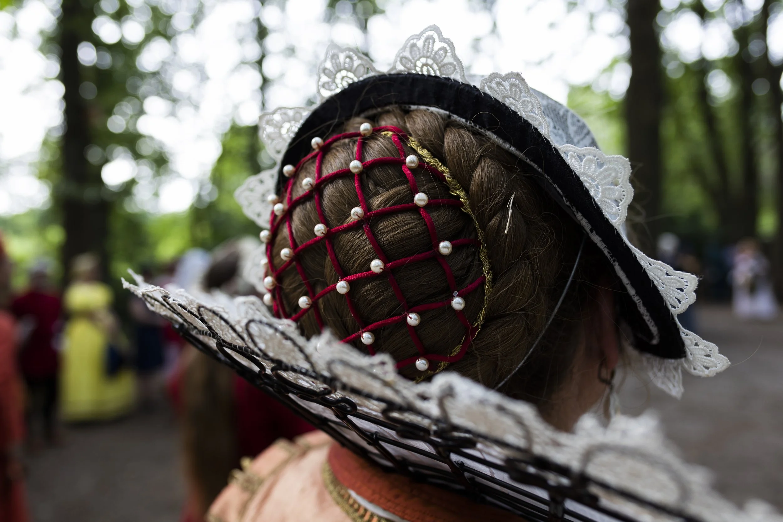 A woman in traditional dress with an ornate, wide-brimmed hat decorated with white lace, pearls, and red ribbon, participating in a cultural or festive event outdoors in a wooded area.