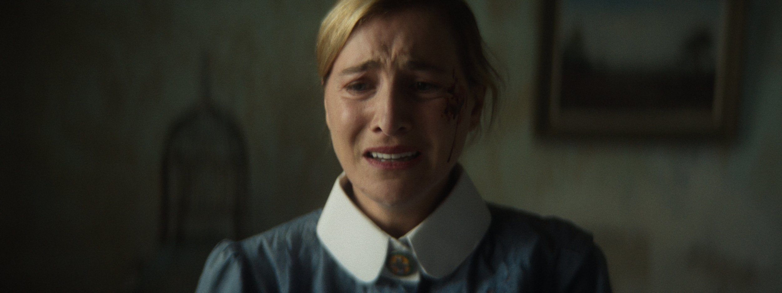 A woman with blonde hair, wearing a blue dress with a white collar, appears distressed with tears and a bloody face, standing indoors with a dark background.