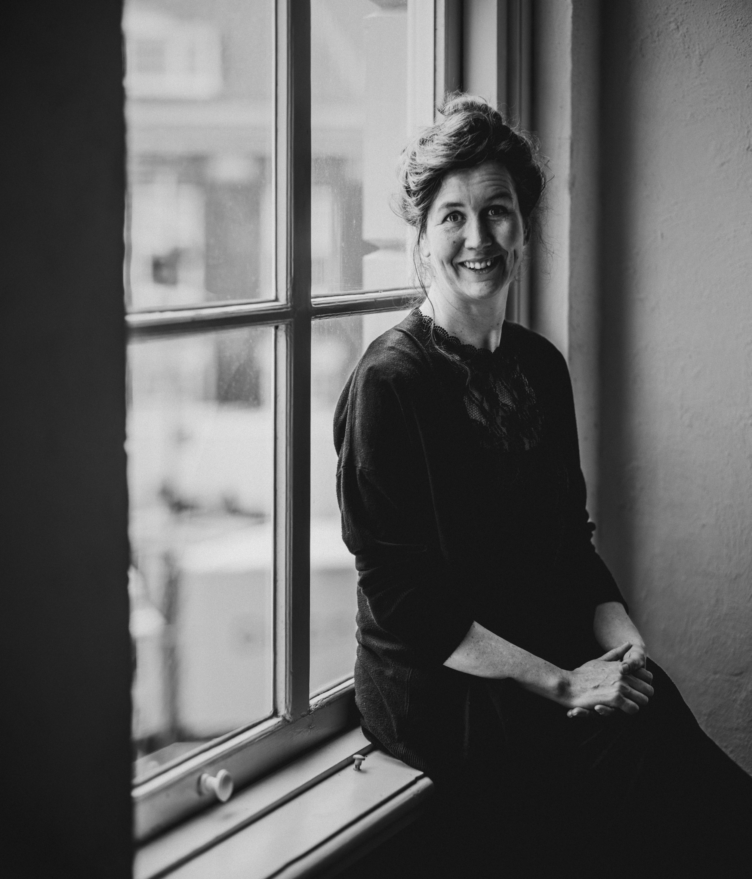Black and white photograph of a woman sitting by a large window, smiling at the camera, with her hands resting on her lap. She has wavy hair and is wearing a dark top with lace detail.