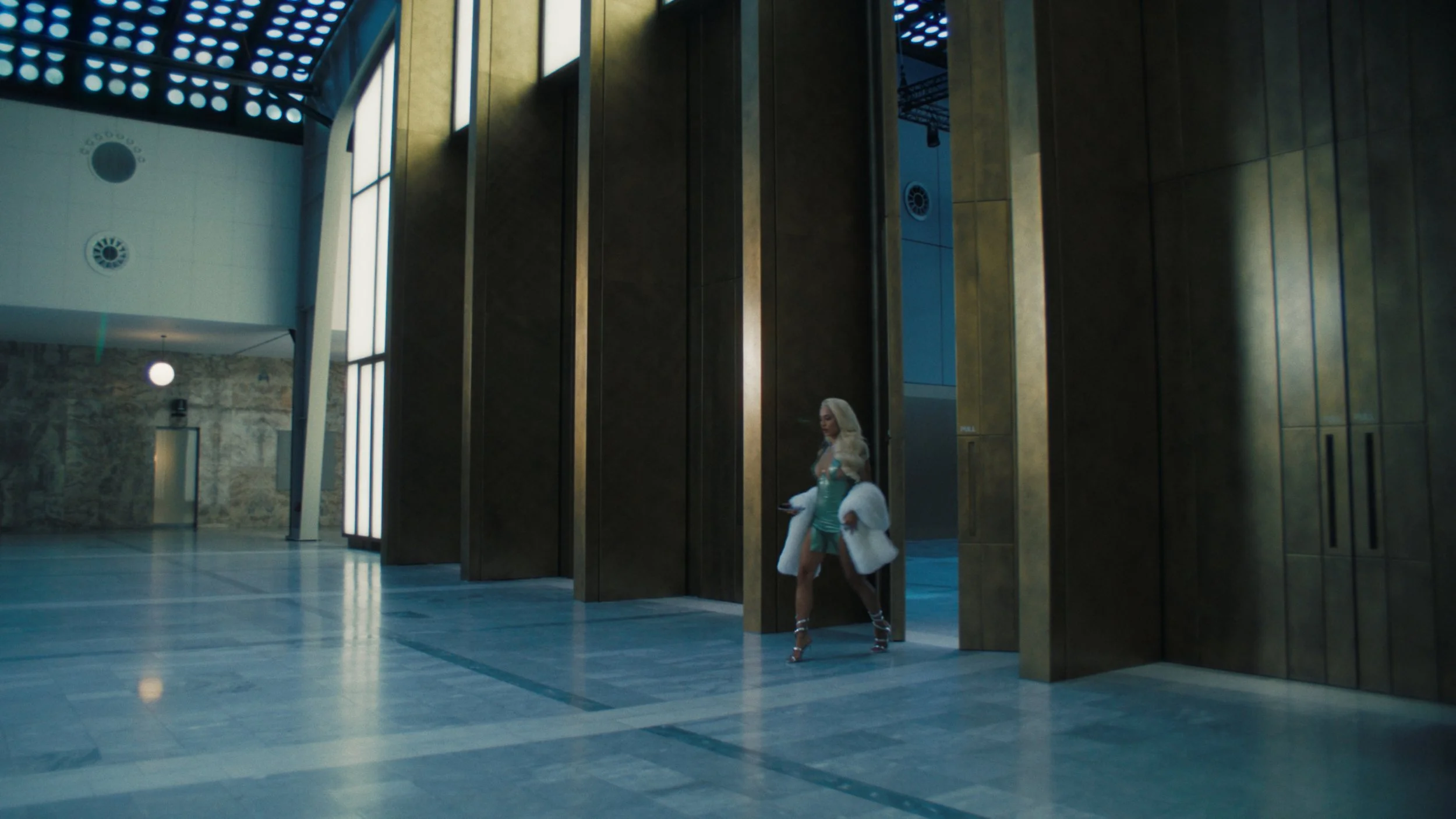 A woman with blonde hair wearing a light blue dress, white fur coat, and high heels standing in an indoor space with tall golden panels, some of the walls have an industrial appearance, and the ceiling features circular light fixtures.